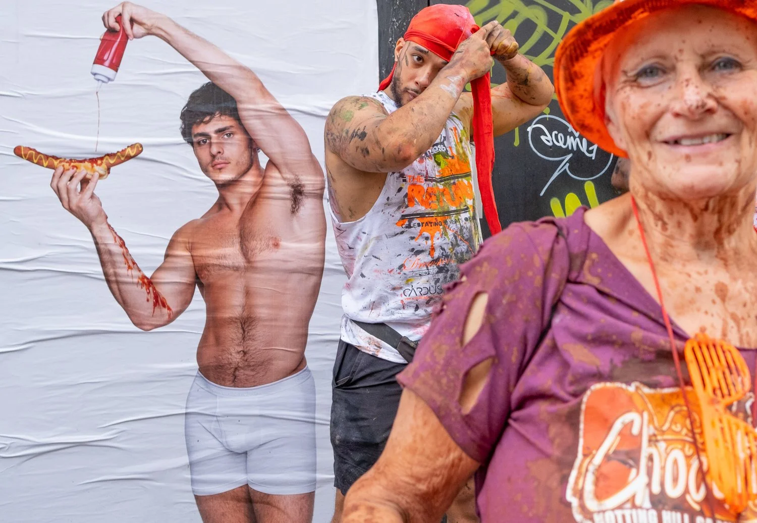 A woman dressed as a hot dog performs at a parade, with a large poster of a shirtless man holding a hot dog in the background, and another person adjusting a red bandana on his head.