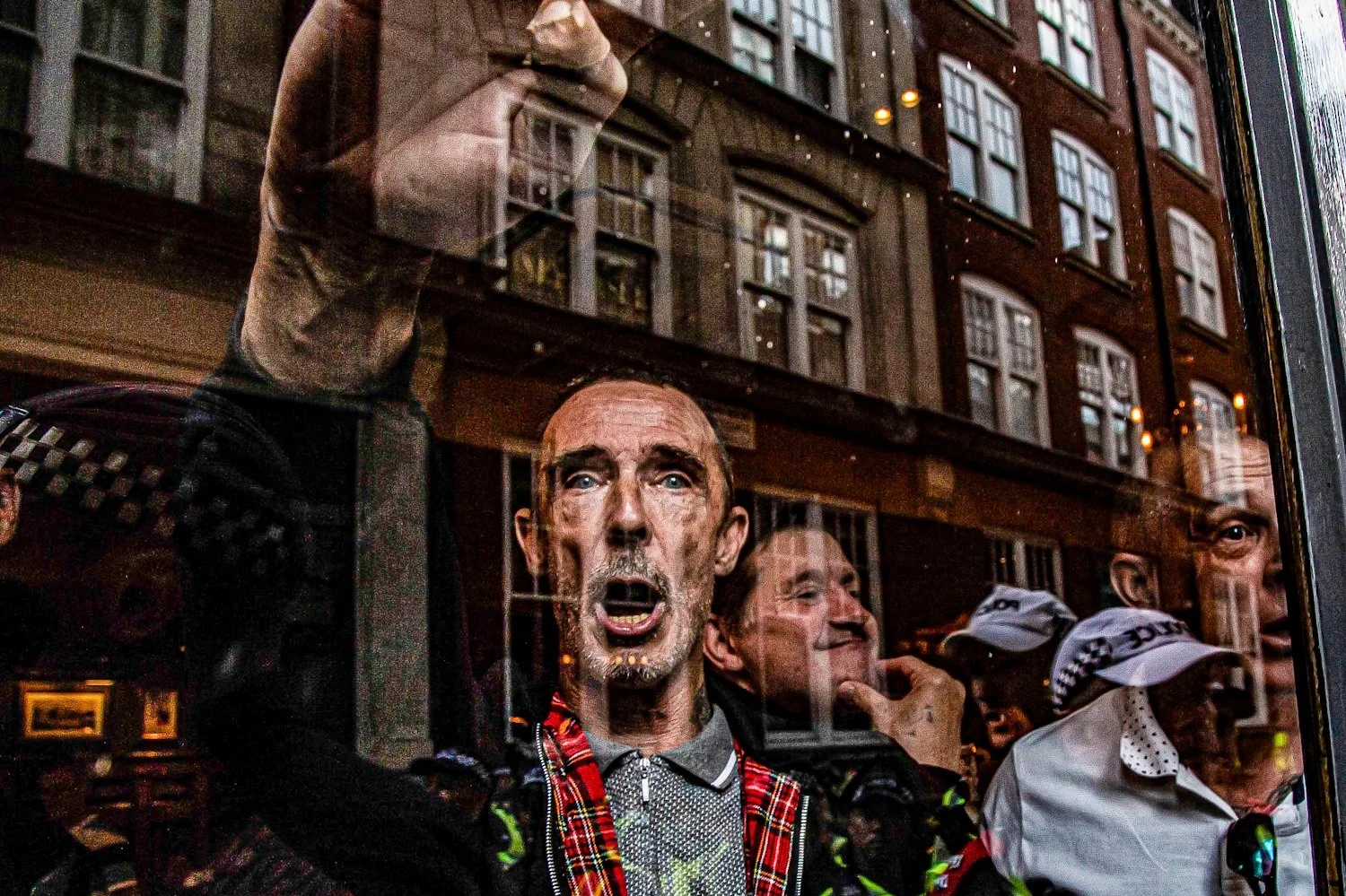 A crowd of people, including firefighters and a man with a tartan scarf, are standing behind a glass window, with some individuals appearing to shout or speak, reflected on a city street with brick buildings and windows.