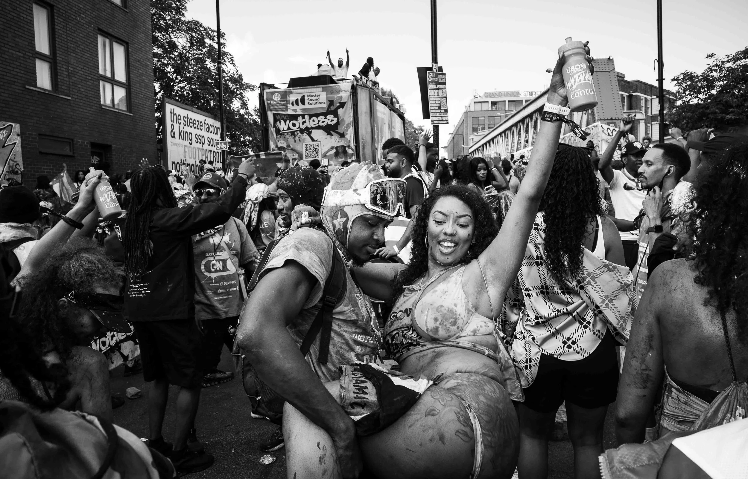 A diverse group of people smiling and dancing at an outdoor event or parade, with some holding drinks and raising their hands, amidst banners, signs, and a cityscape background.