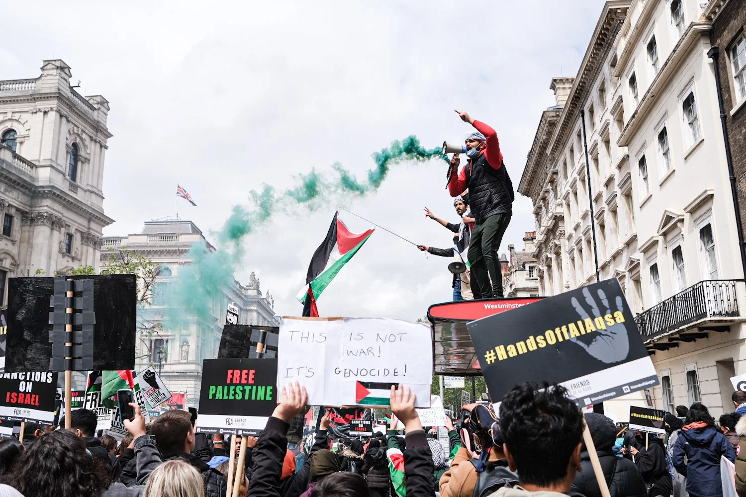 Protest in a city street with a crowd holding signs and banners advocating for Palestine. A man on a float with a megaphone and Palestinian flags is speaking or leading chants, and some protesters are raising their fists. Signs include messages like 