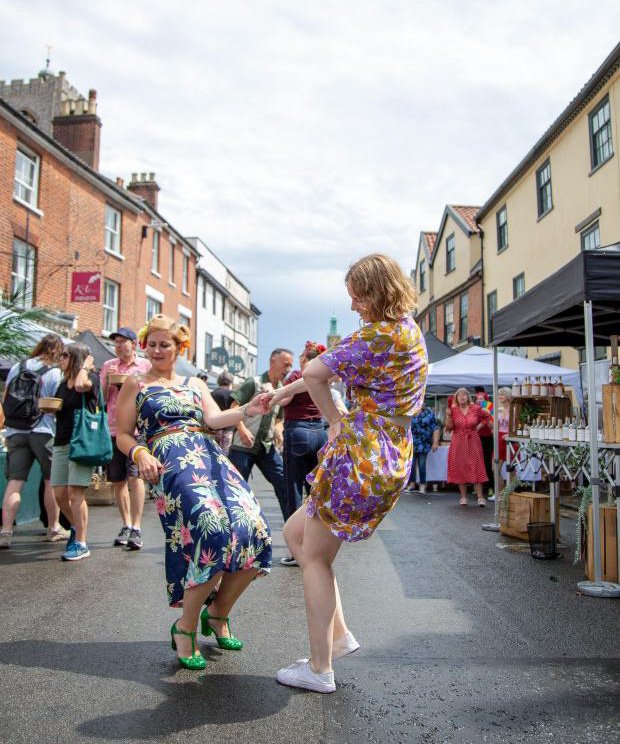 Two women dancing in the street during a festival, with booths and crowd in the background, overcast sky.