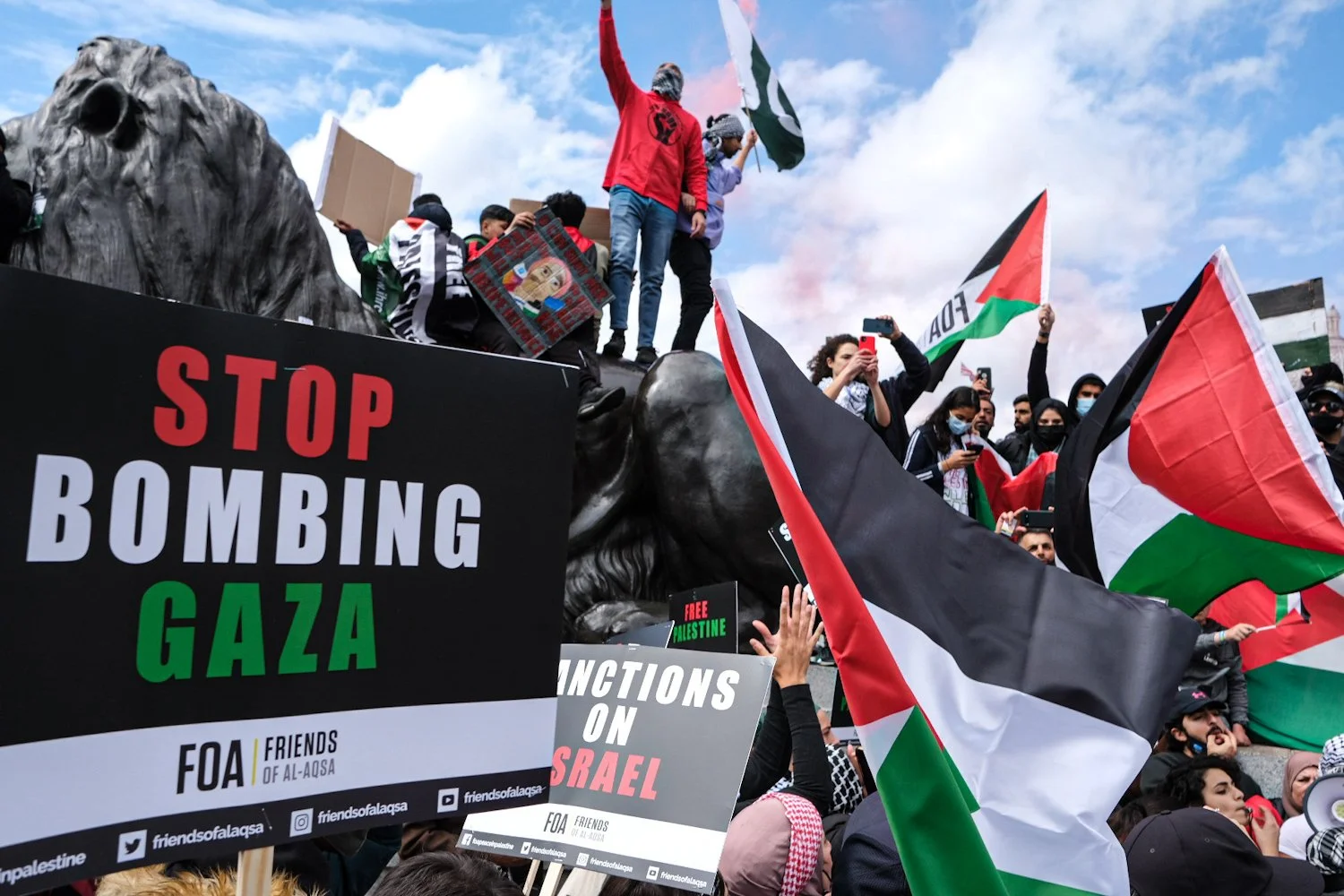 Protesters holding signs and Palestinian flags, with a large lion sculpture and a blue sky in the background. Signs include messages like "Stop bombing Gaza" and "Actions on Israel."