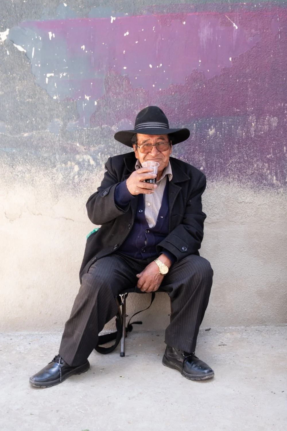 An elderly man sitting on a small stool against a colorful, textured wall, wearing a black wide-brimmed hat, glasses, a black jacket, checkered shirt, pinstripe pants, black shoes, and gold watch, holding a glass of dark beverage.