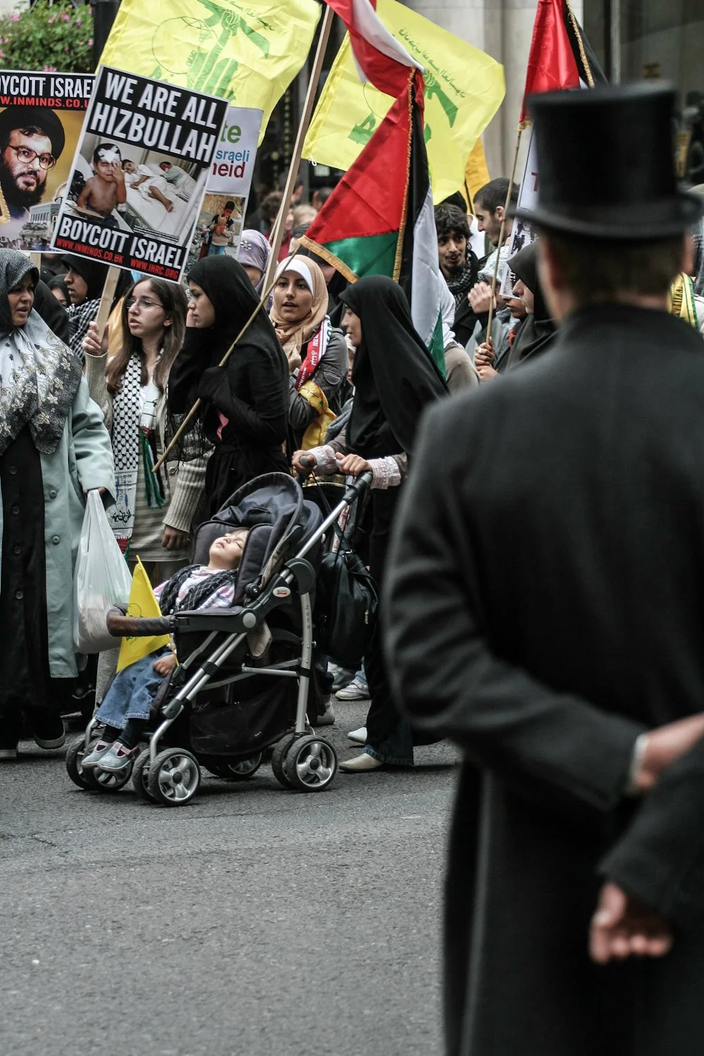 A crowd of people participating in a protest or demonstration, holding signs and flags supporting Palestine, with some individuals wearing traditional clothing and a person pushing a young girl in a stroller.