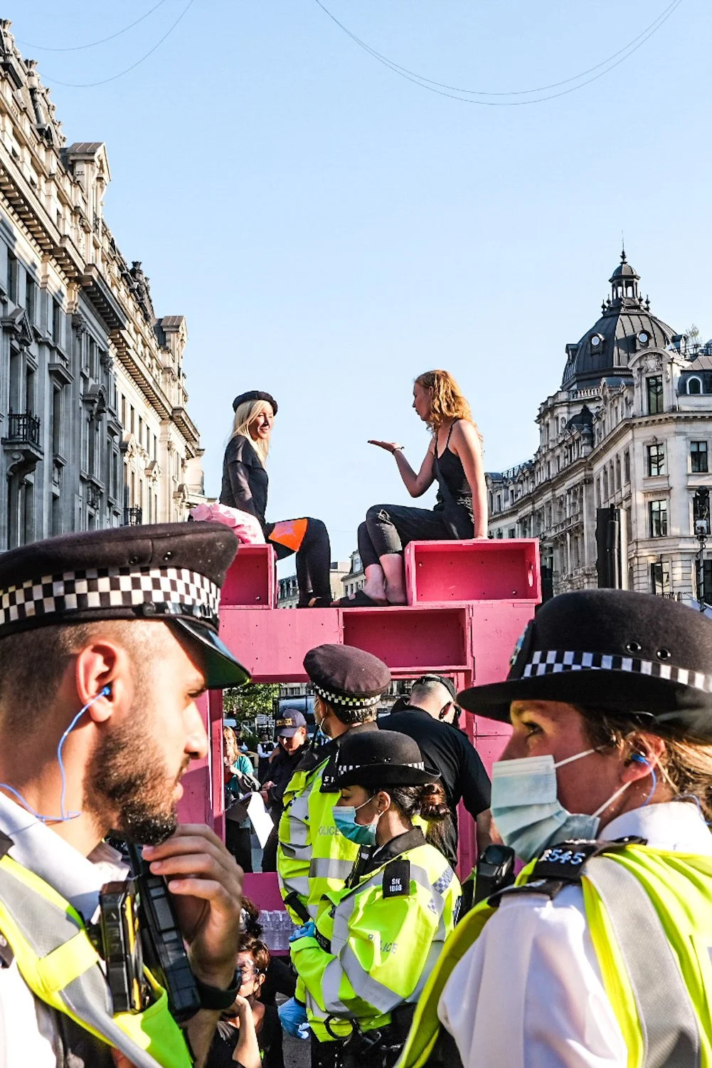 Two women sitting on a pink float protesting, with police officers and crowd in the foreground, in an urban city street.