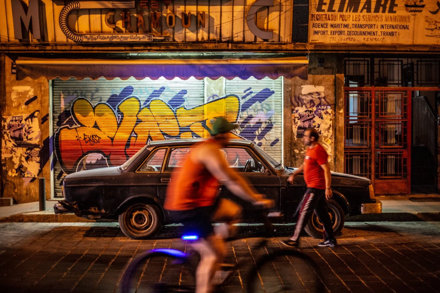 A nighttime street scene featuring a black vintage car parked in front of a graffiti-covered storefront with metal shutters. Two men in red shirts are standing and talking near the car, and a person is riding a bicycle in motion in the foreground. Th