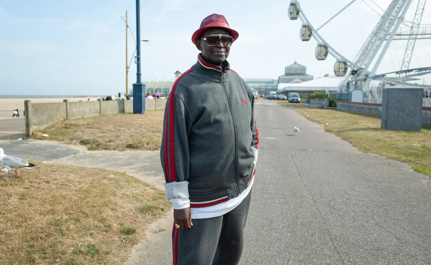 A man wearing a red hat, sunglasses, and a gray and red track suit standing on a promenade near a Ferris wheel at a beachside location.