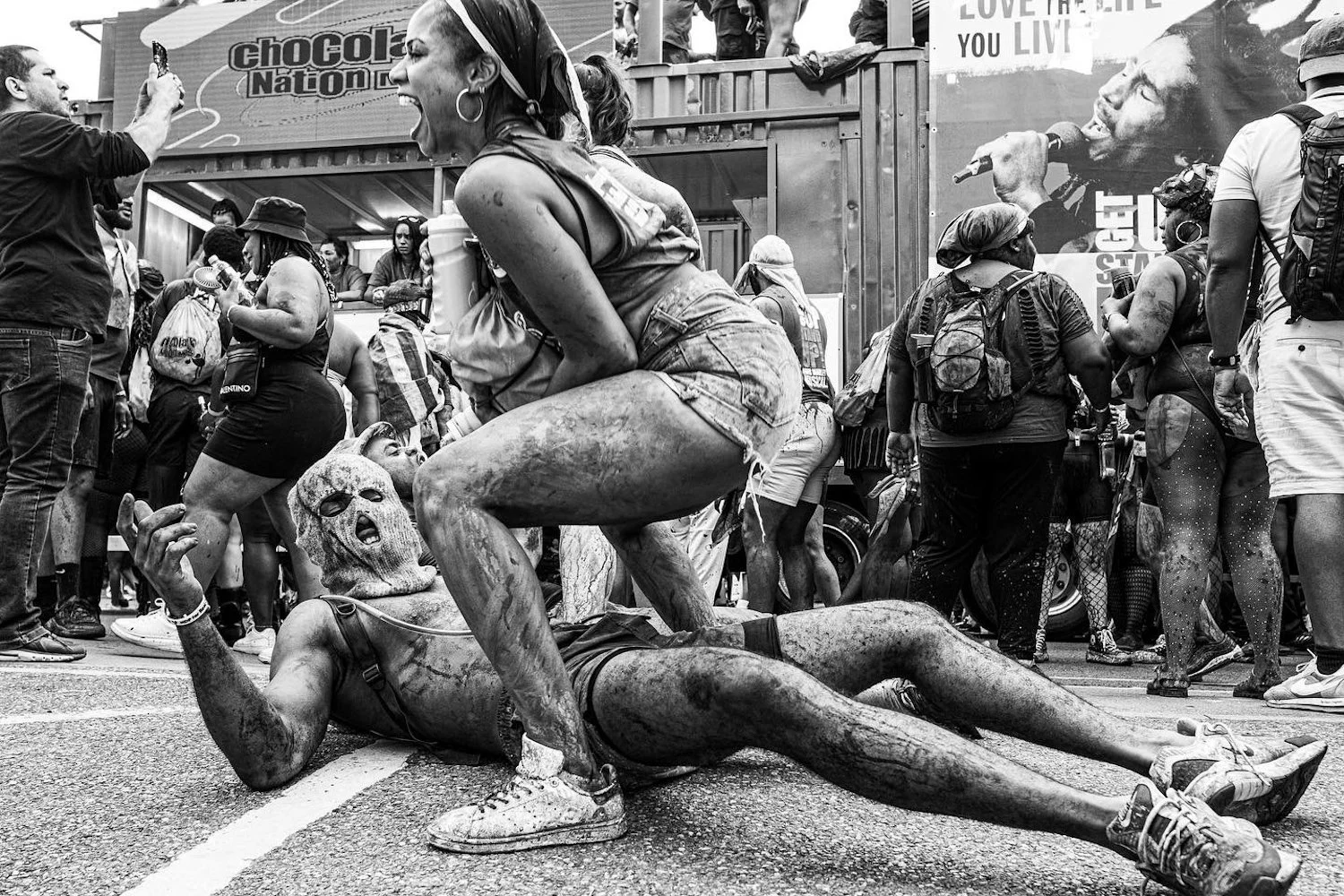 Two women with mud-covered bodies engaging in a playful fight during a protest or event, surrounded by a crowd of people with backpacks and casual clothing, in an urban setting.