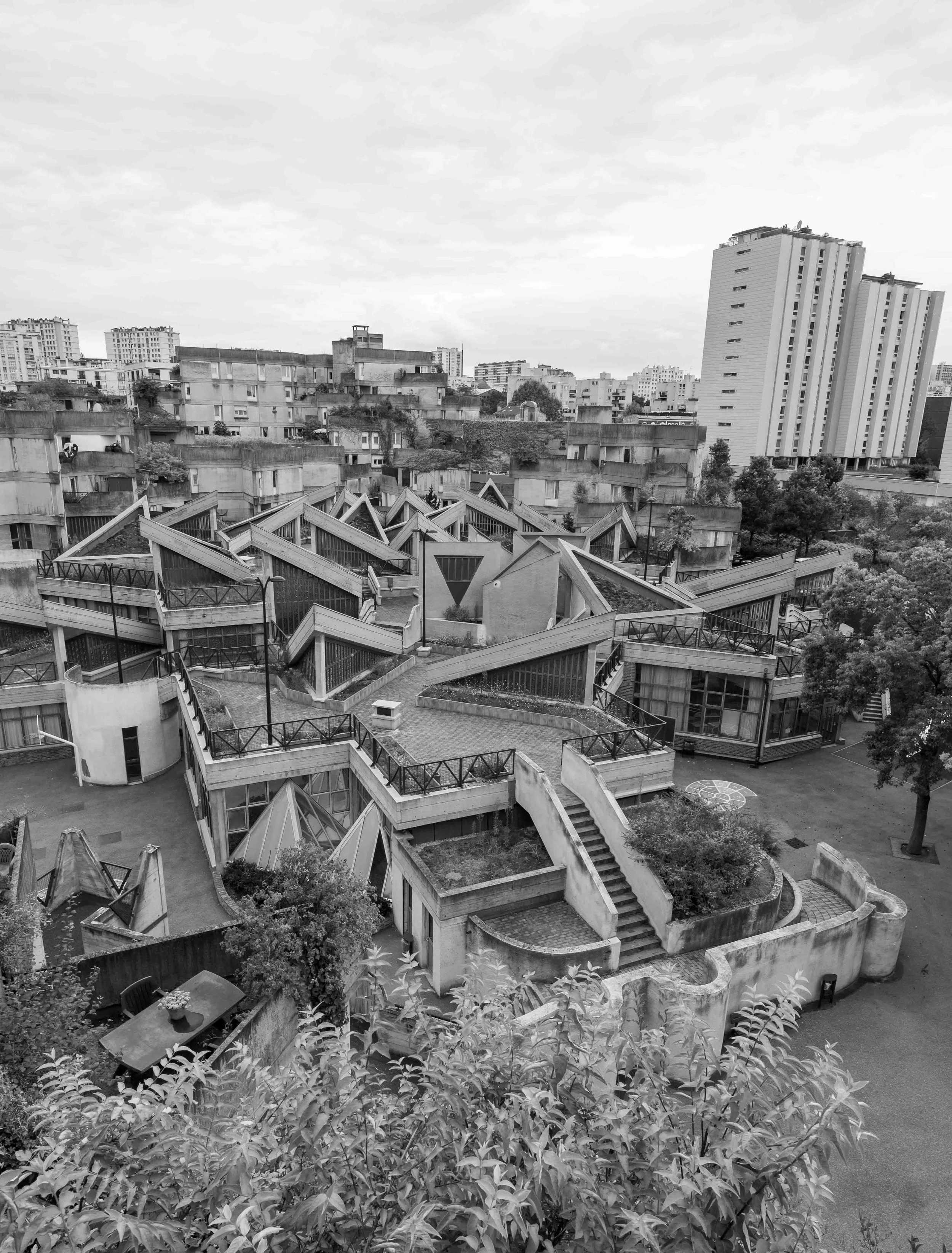 A cityscape with modern and older buildings, features a complex architectural structure with stairs and walkways, trees, and residential high-rises in the background, in black and white.