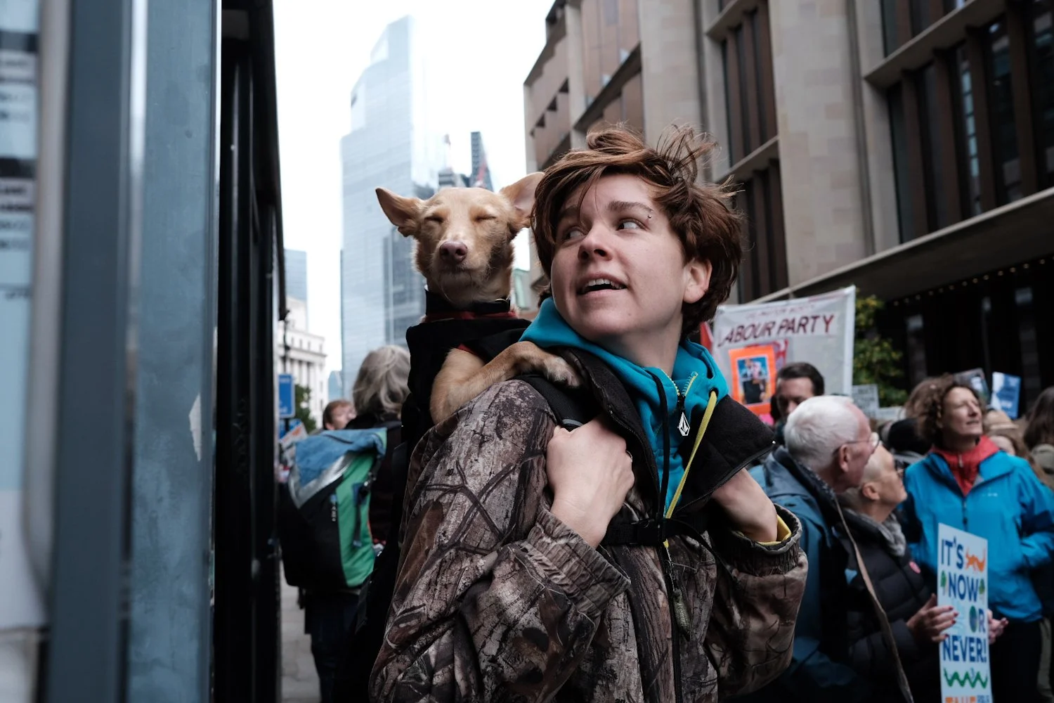 A young person carrying a small dog on their shoulder during a protest or rally in an urban setting with tall buildings and a crowd of people holding signs.