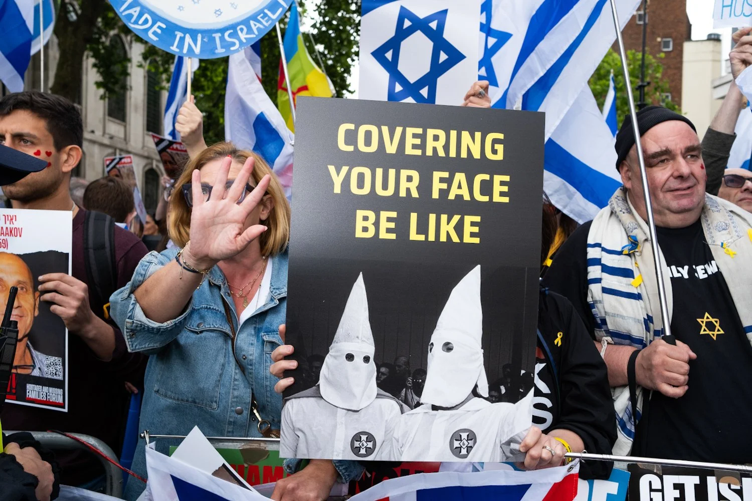 Protesters holding flags and signs, including one that reads 'COVERING YOUR FACE BE LIKE' with an image of two people in hooded costumes with eye holes, at a public demonstration.