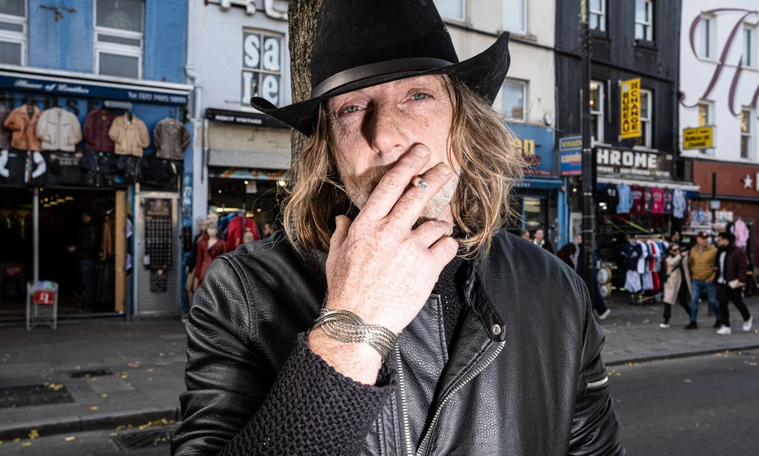 A man with long, wavy hair, wearing a black leather jacket and a black cowboy hat, standing on a city street with shops and pedestrians in the background.