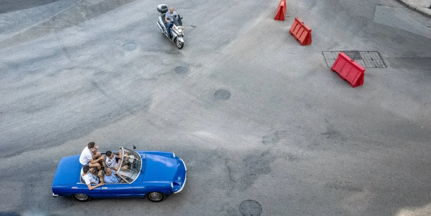 An aerial view of a blue convertible car with four people sitting inside and one person in a striped shirt standing nearby. A person riding a scooter is passing by on the street, which is marked with orange traffic barriers and manhole covers.
