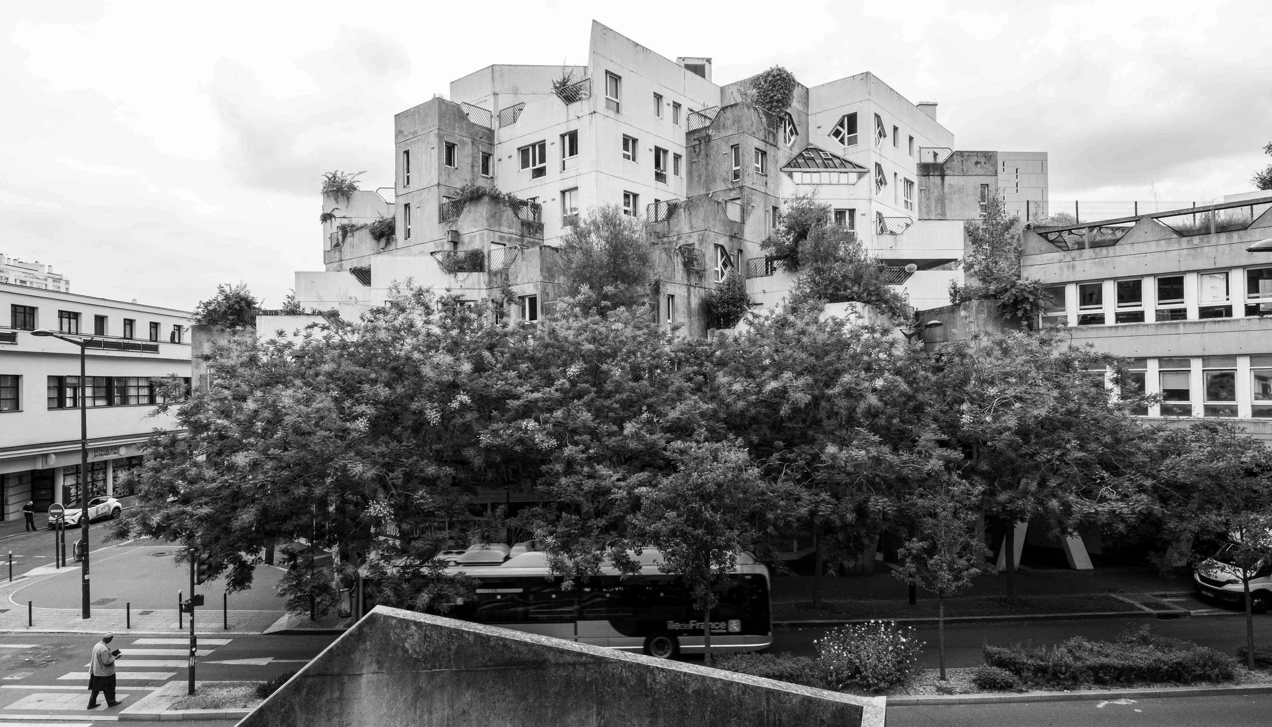 Black and white photo of a modern, multi-story apartment building with irregular windows and balconies, surrounded by trees and urban street scene.