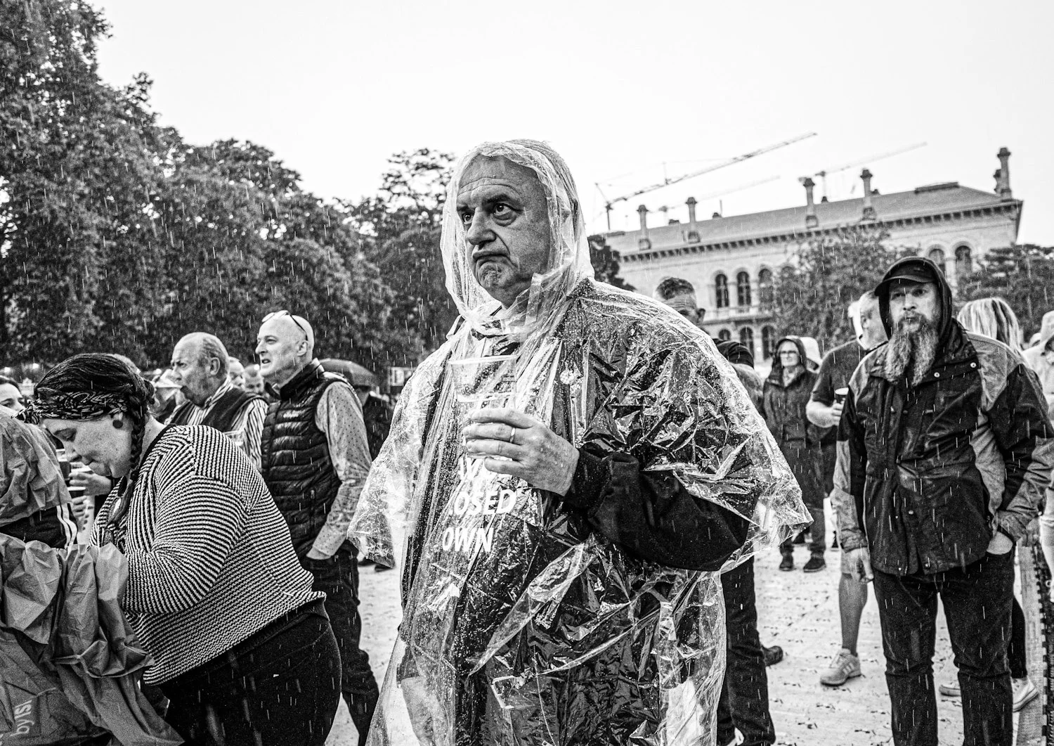 A group of people gathered outdoors in the rain, with one man wearing a clear rain poncho and carrying a sign that says "Closed Own." The man appears to be angry or upset, and the background shows trees and a building under construction.