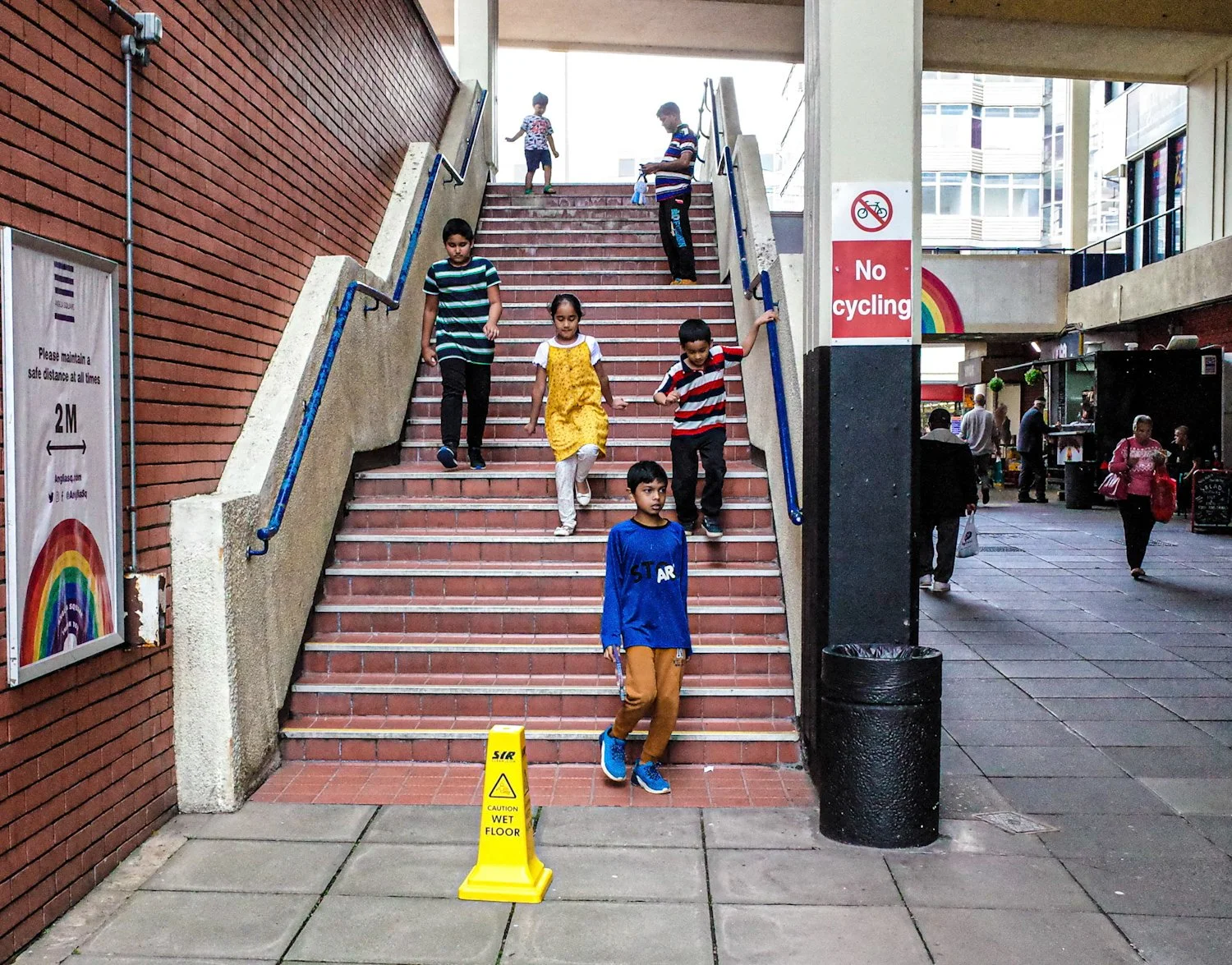 A group of children descending indoor stairs in a shopping mall or public space. Some adults are visible in the background, along with a no cycling sign and a colorful rainbow decoration.