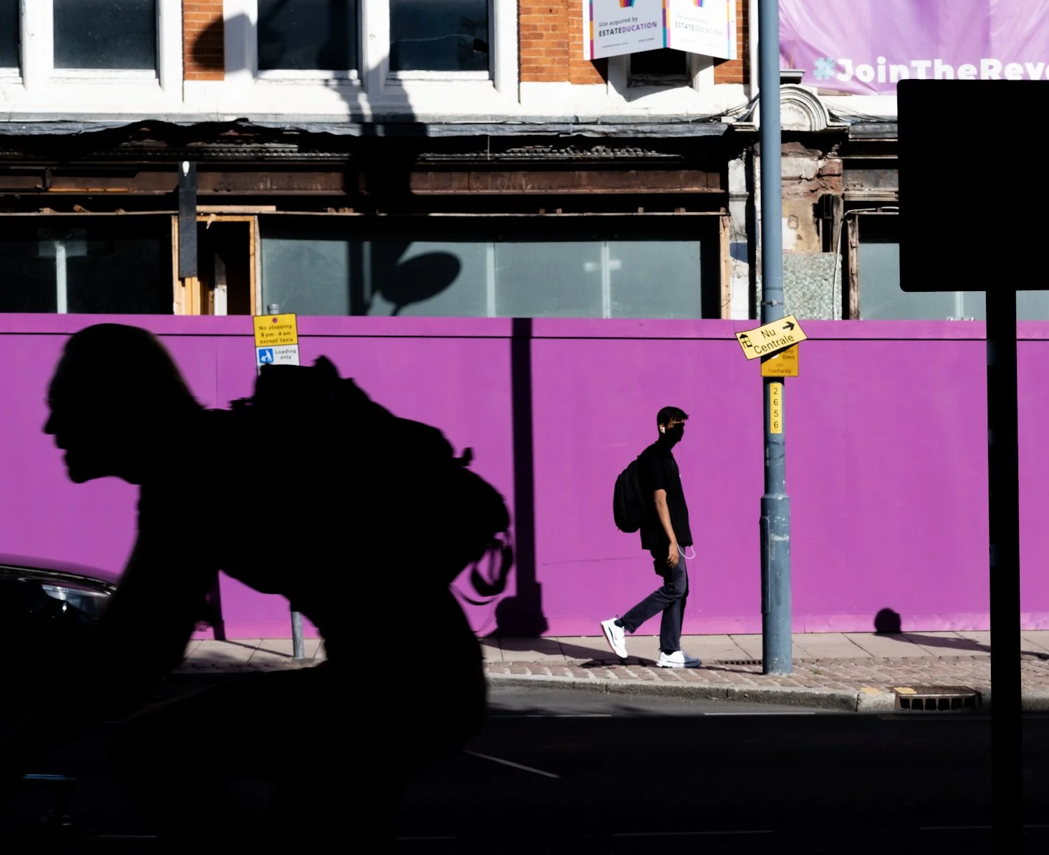 A person walking on the sidewalk wearing a black mask, black t-shirt, gray pants, white sneakers, and carrying a black backpack. A silhouette of a motorcyclist rides by in the foreground. The background features a tall purple wall, a pole with street