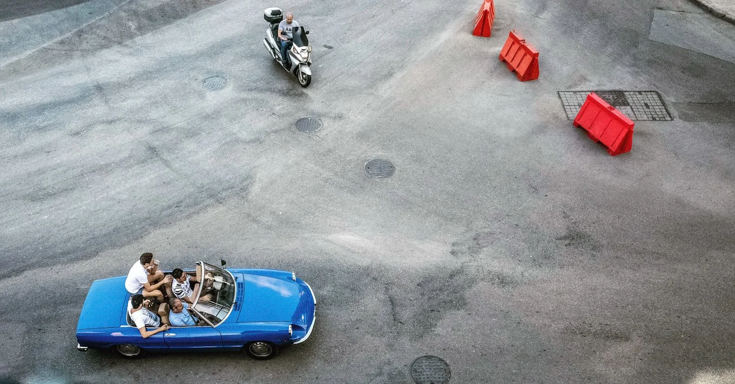An aerial view of a blue convertible car with five people inside and a motorcyclist passing by on the street. There are orange barricades and a drain grate on the pavement.