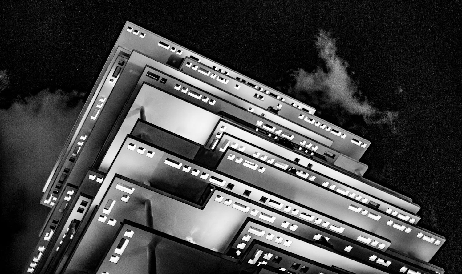 Low-angle view of a modern multi-story building at night with illuminated windows and a cloudy sky in the background