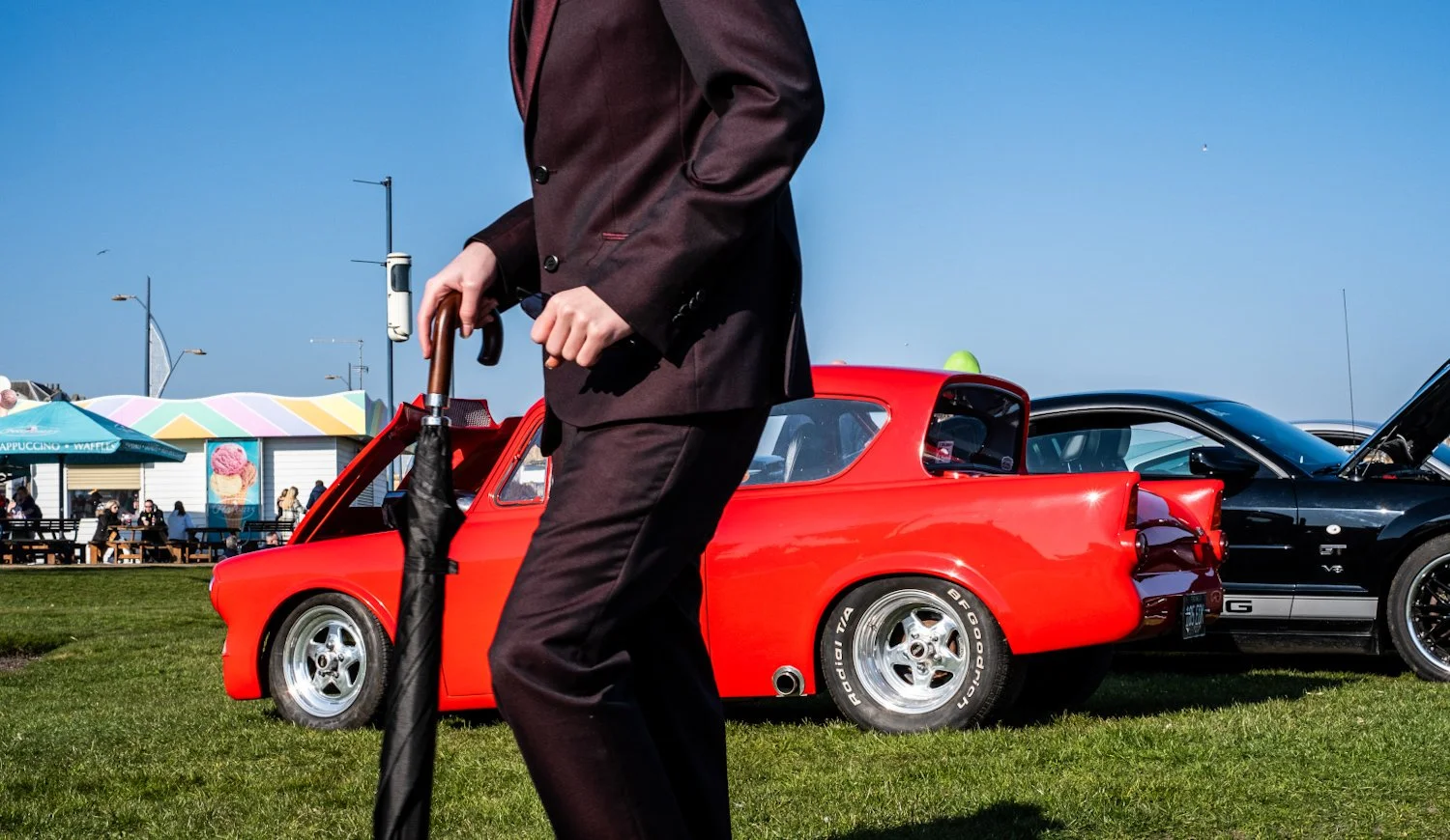 A man in a black suit standing outdoors on a grassy area, holding a closed black umbrella. Behind him are two cars, one bright red and the other black, parked next to each other. In the background, there are food stalls and people sitting at benches,