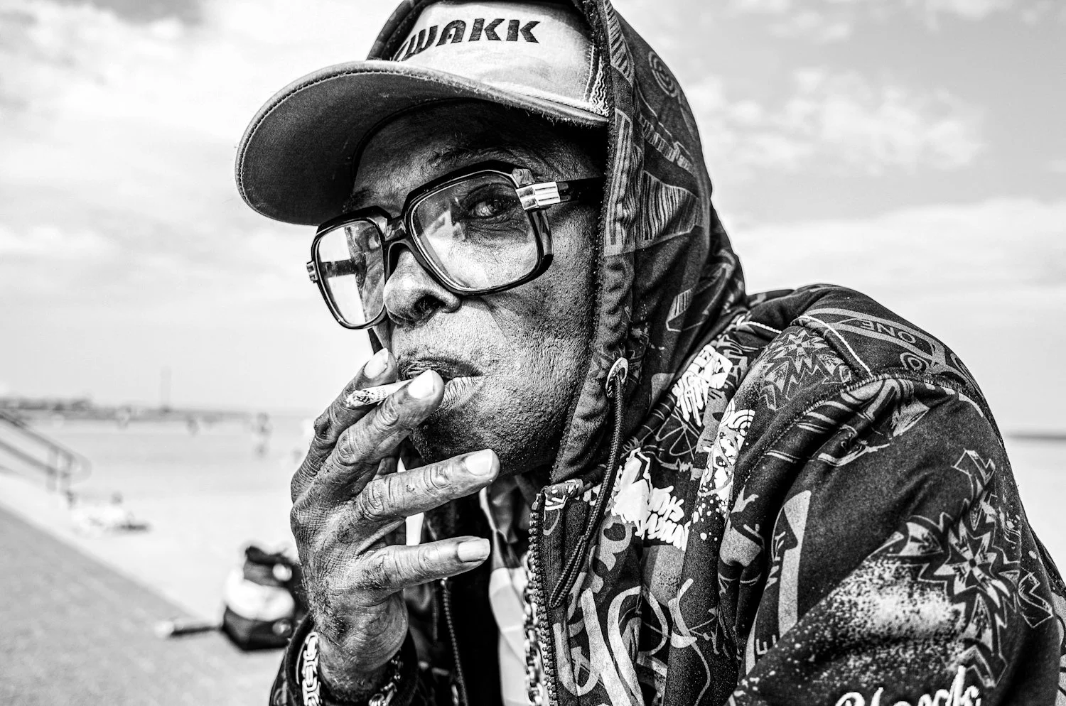 A black-and-white photo of a man wearing a baseball cap, large glasses, and patterned clothing, smoking a cigarette near a body of water.