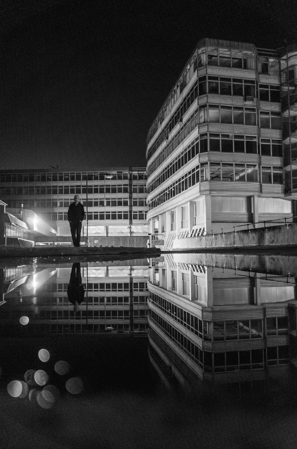 Nighttime cityscape with modern multi-story building, a person walking, and reflections in a body of water.