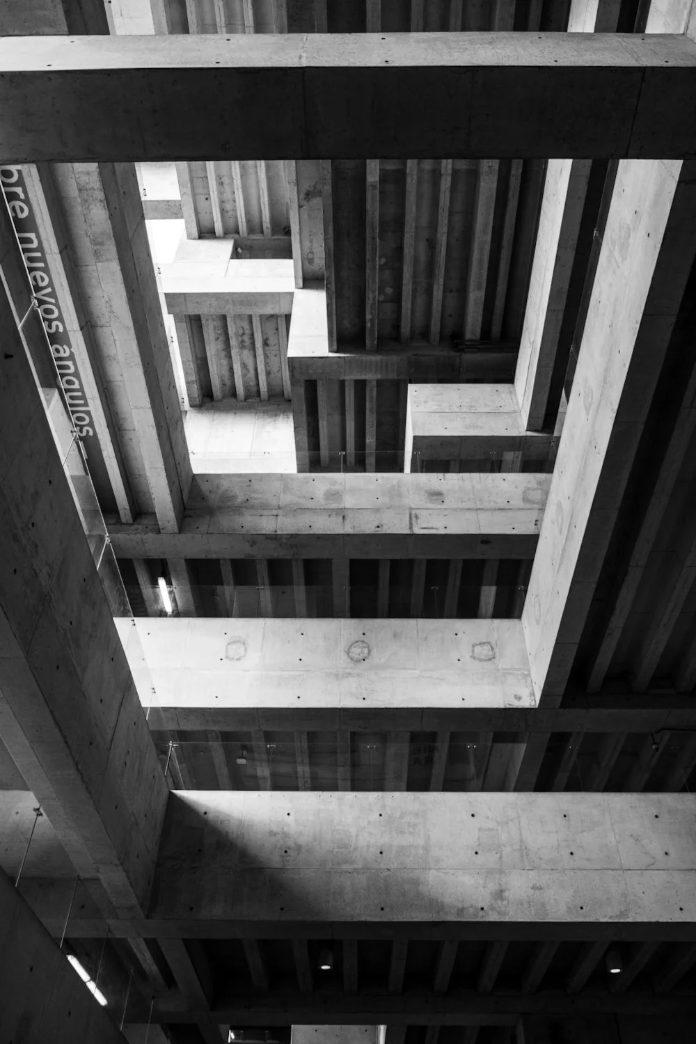 Black and white photo of a modern building's interior showing multiple open floors with concrete beams and railings, looking up towards the ceiling.