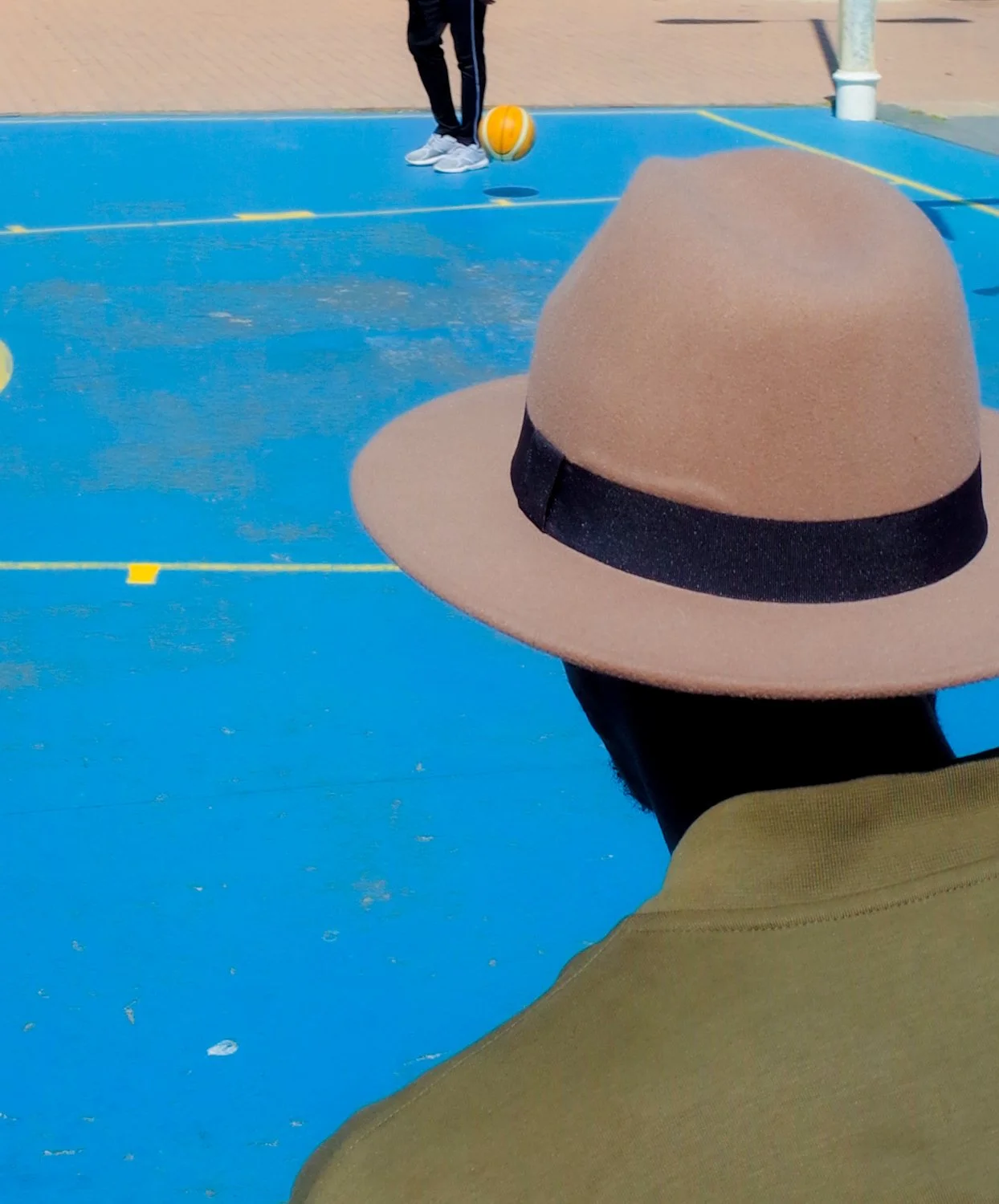 Person wearing a pink wide-brimmed hat and yellow hoodie, facing away, watching a basketball game on an outdoor blue court with yellow lines. Two players are visible, one dribbling the ball.