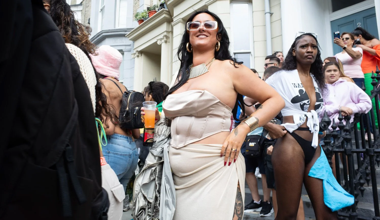 Group of people outdoors at a festive event, with a woman in the center wearing a beige dress, glasses, gold jewelry, holding a drink, and smiling.