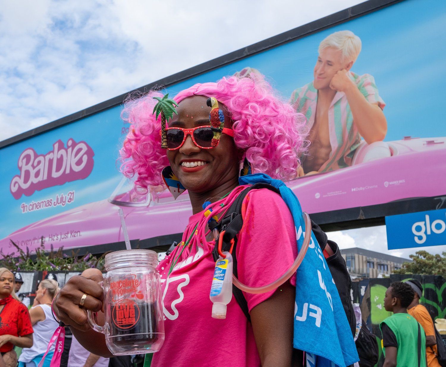 A smiling woman with pink curly hair wearing colorful glasses with a parrot and palm tree design, pink shirt, backpack, and holding a clear mug, standing in front of a billboard advertising Barbie movie.