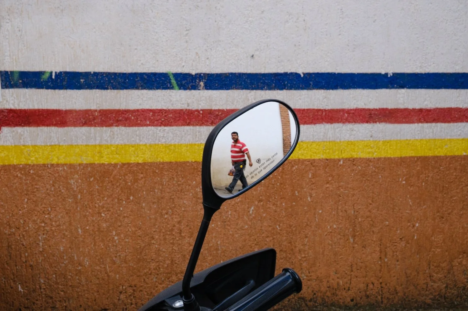 A motorcycle's side mirror shows a man in a red and white striped shirt walking past a wall with horizontal colored stripes in blue, red, yellow, and brown.