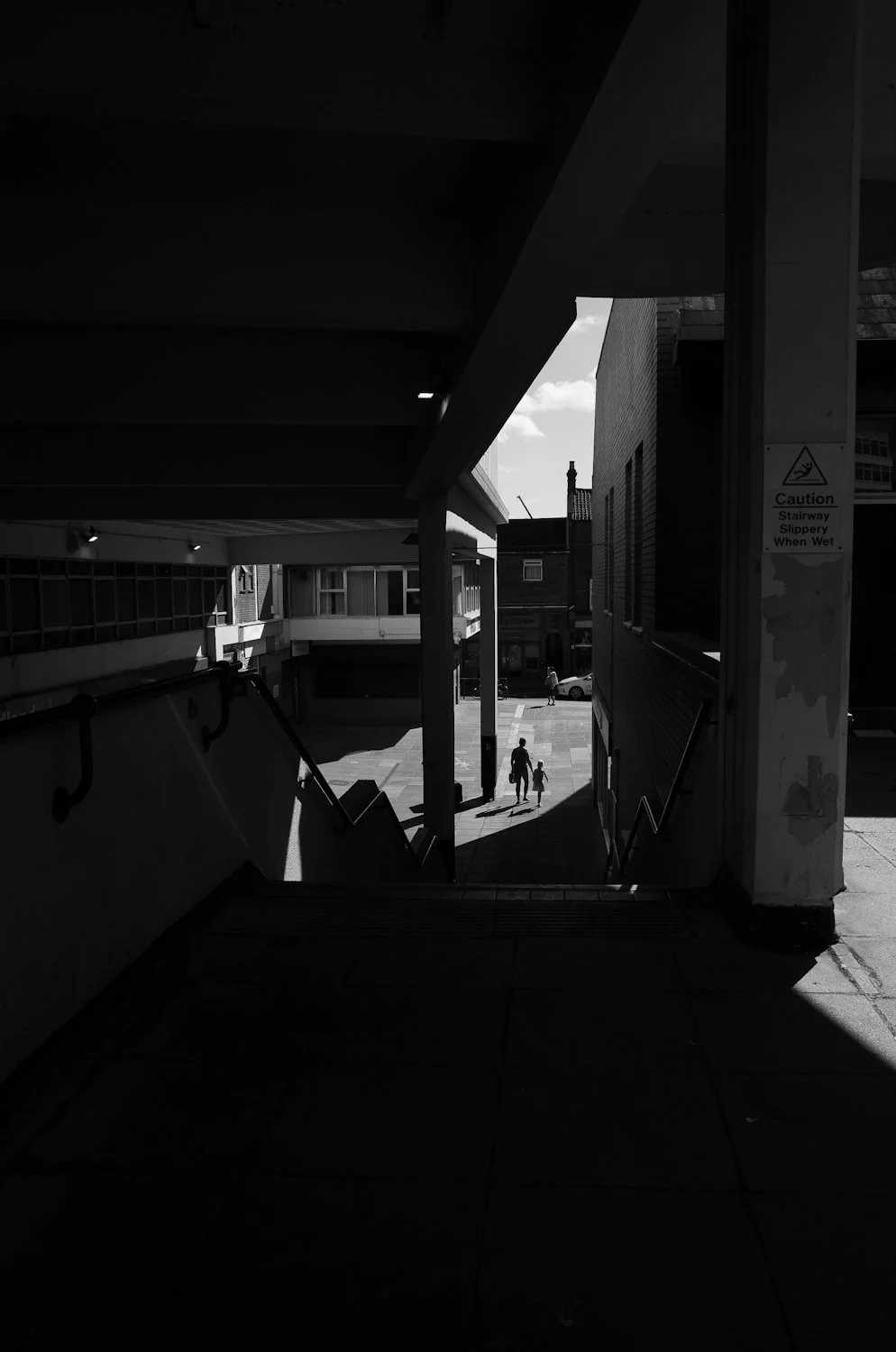 City scene viewed from a dark underpass showing people walking outside in sunlight, with a sign warning of a slippery stairway when wet.
