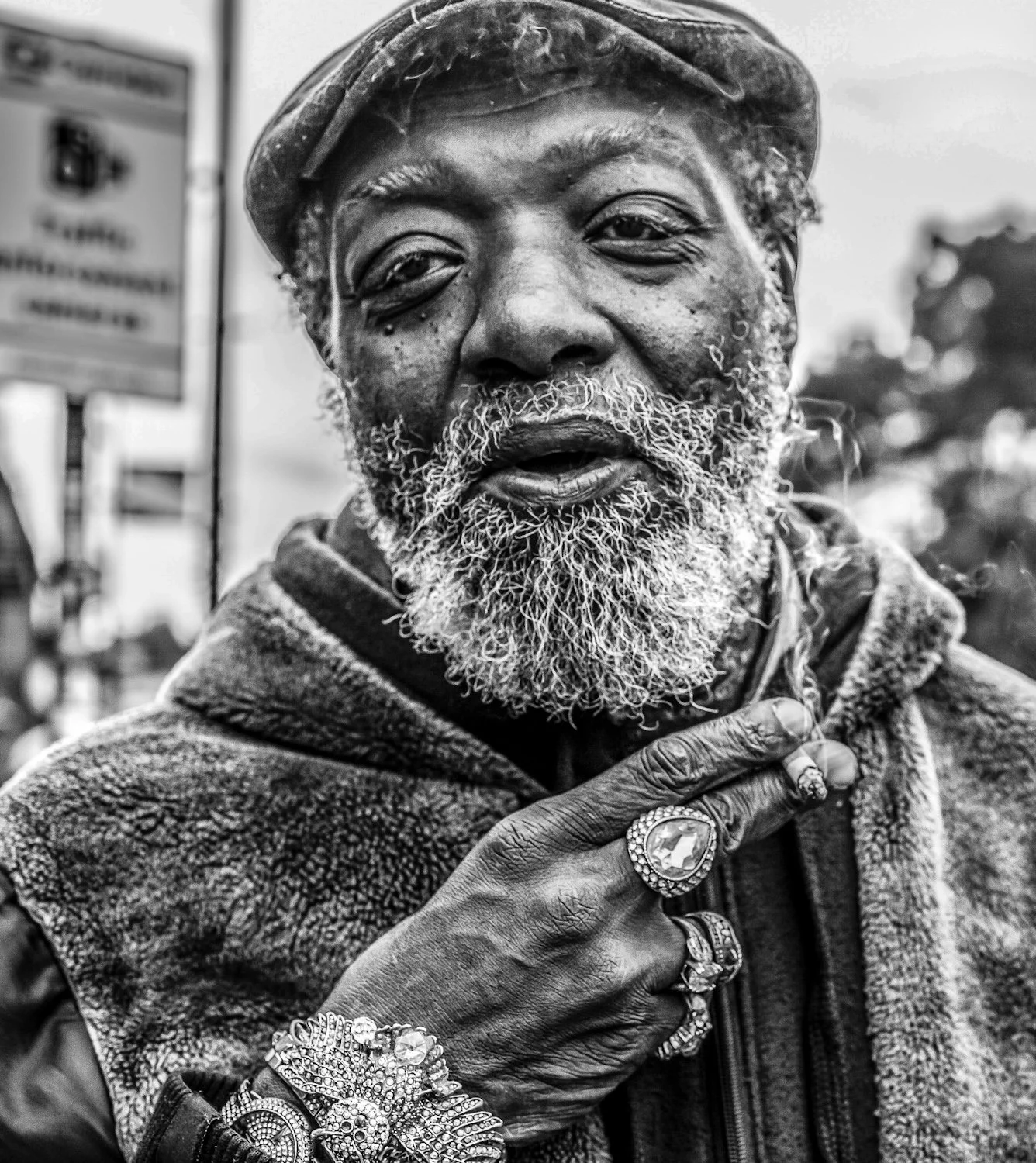 An elderly Black man with a beard, wearing a hat and jewelry, holding his hand to his chest, in an outdoor setting.