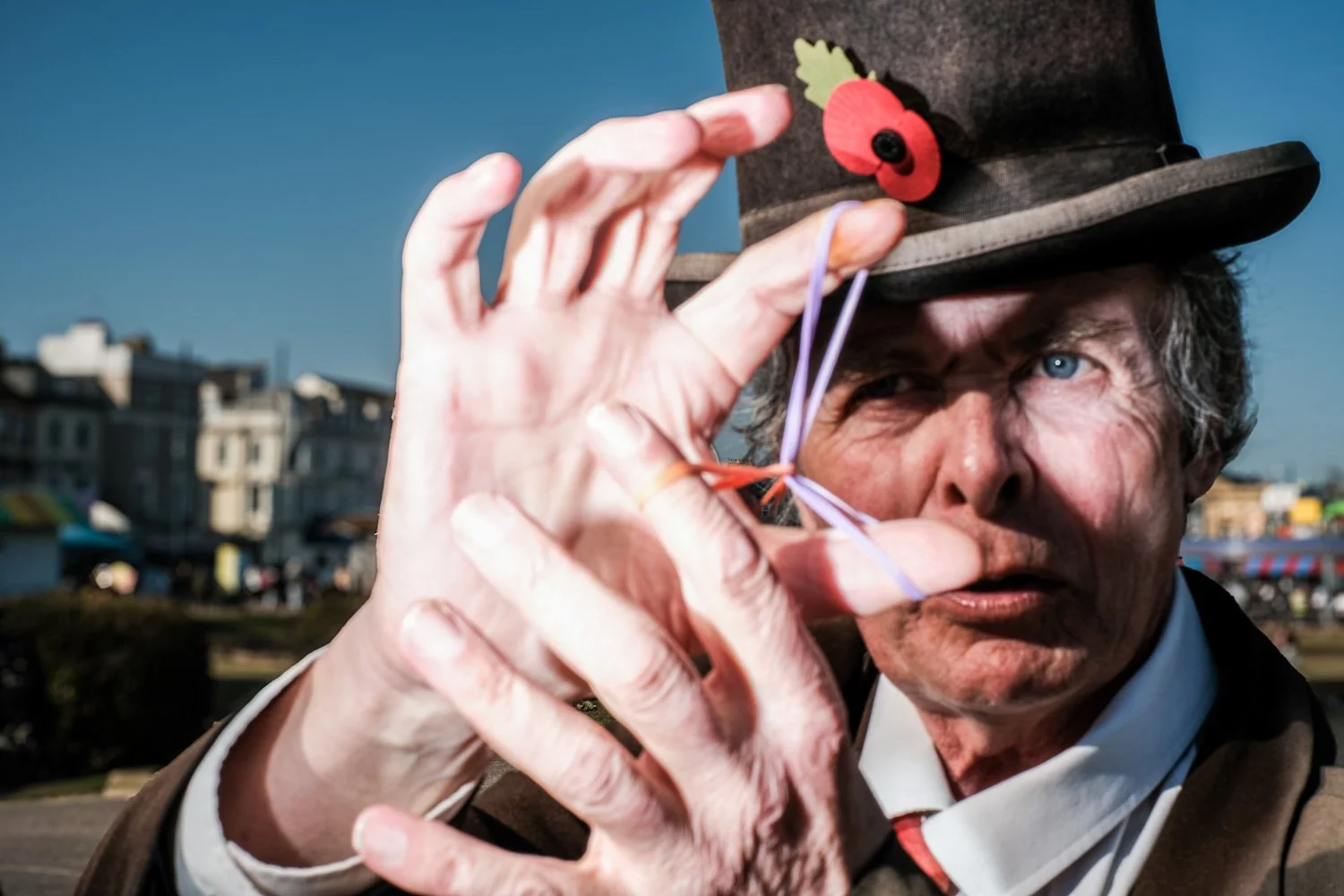A man wearing a tall, dark hat with a festive red and green decoration, holding his hand close to his face, making an 'OK' gesture, outdoors with a cityscape in the background.