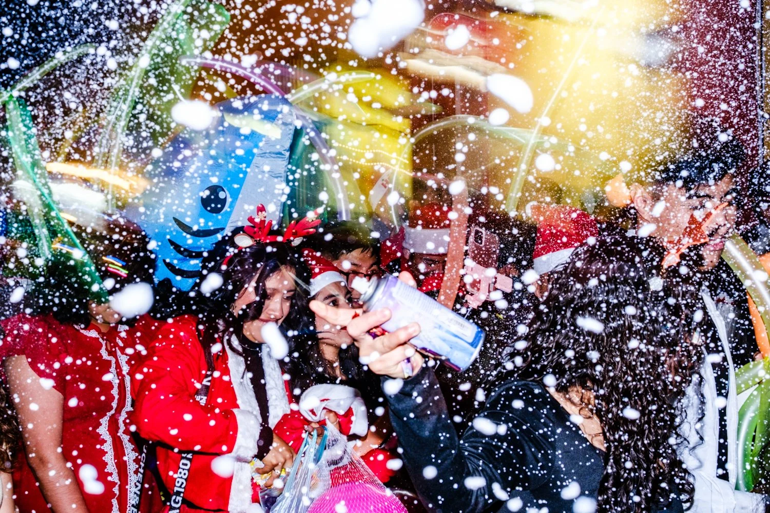 Group of people celebrating Christmas with fake snow, wearing festive outfits, including Christmas hats and reindeer antlers, with colorful decorations in the background.
