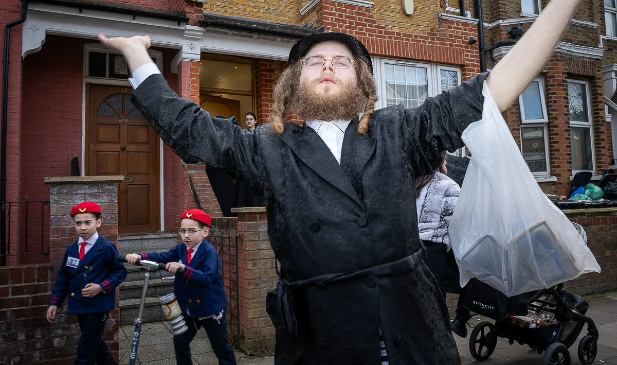 Man with glasses and a beard, wearing a black coat and a hat, standing with arms outstretched outside a brick residential building.