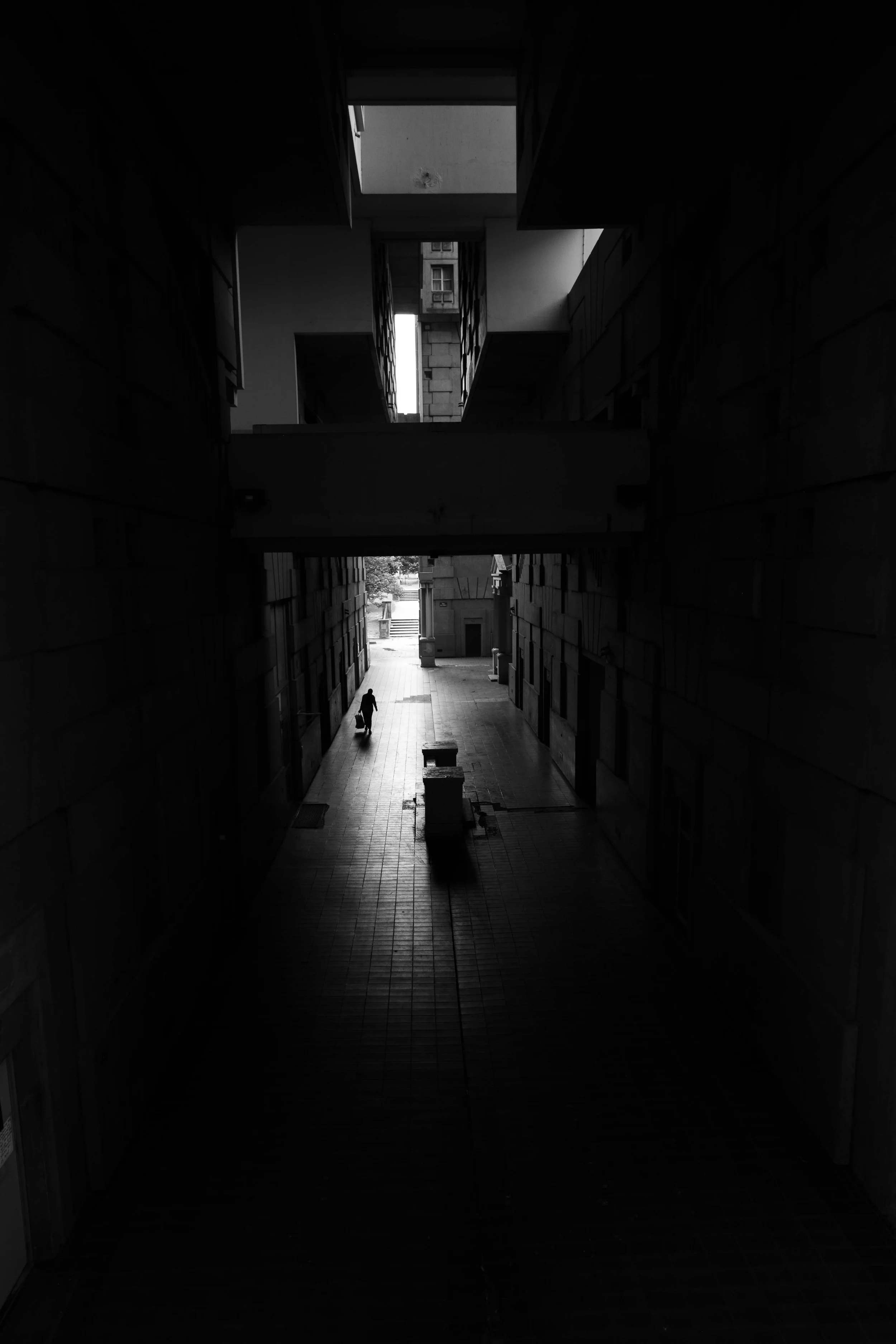 A person walking with a suitcase in a dark corridor leading to a brighter outside area.
