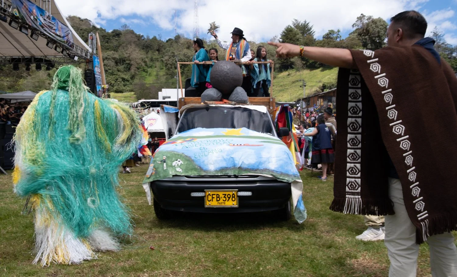 People in traditional clothing participating in a cultural event surrounding a decorated vehicle with artwork and a license plate reading 'CFB-398 PASTO', with an outdoor stage and crowd in the background.