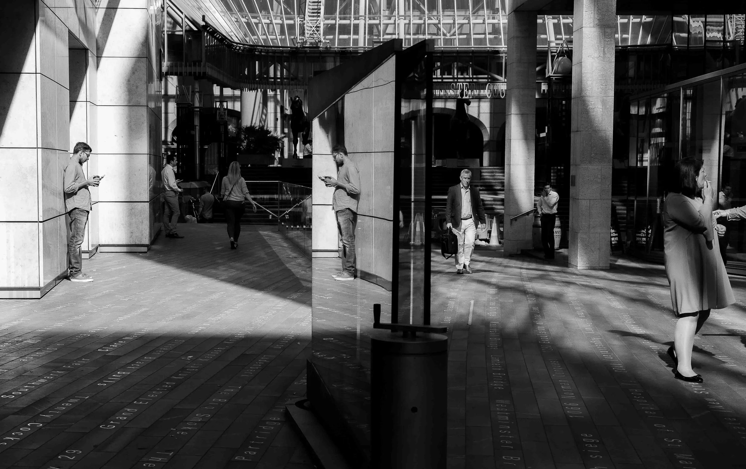 Black and white photo of a busy urban scene with people walking and standing, some on their phones, in front of a modern building with glass walls, columns, and sculptures. A glass door reflects the street scene outside.
