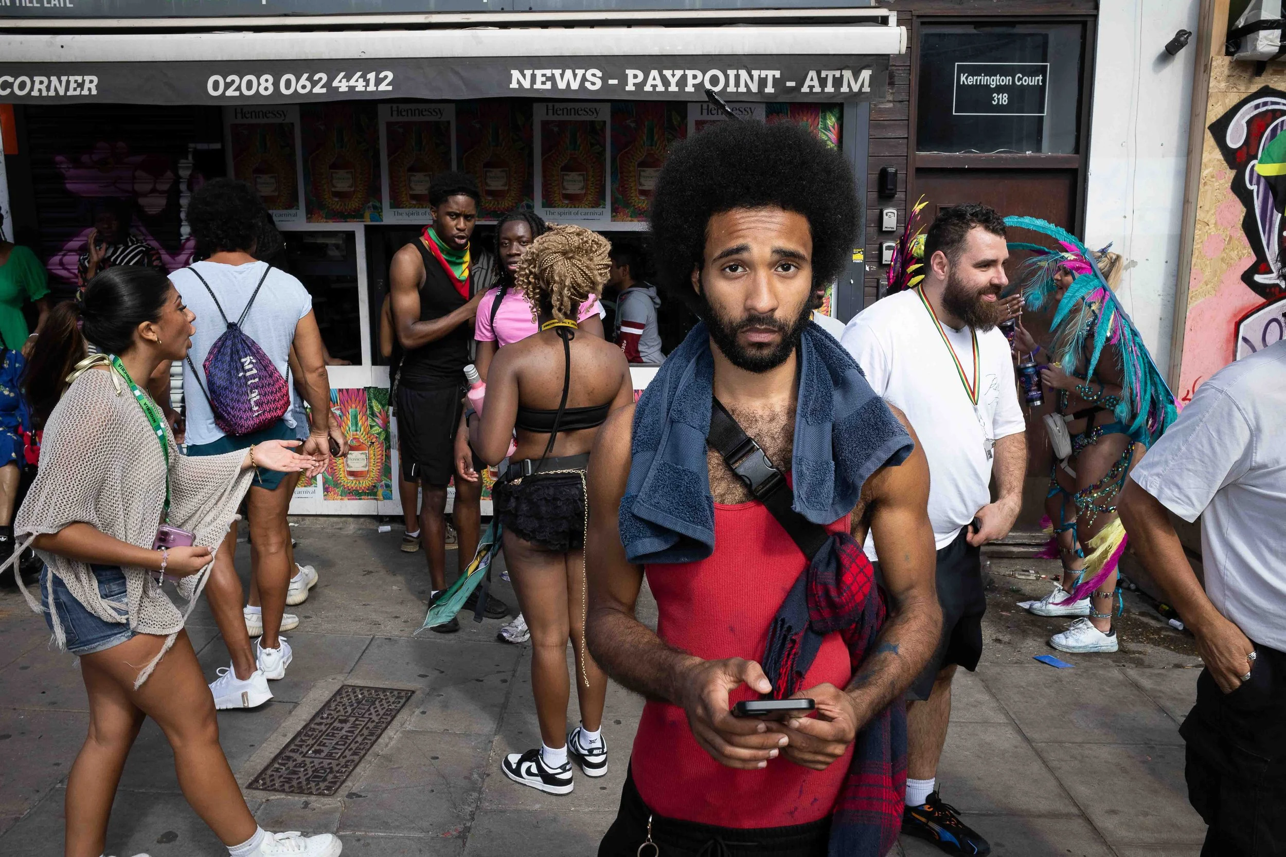 A group of diverse people gathered outside a building, with one man in the foreground wearing a red shirt and a black and red flannel shirt, holding a phone, with several others in the background, some dressed in colorful or festival attire.
