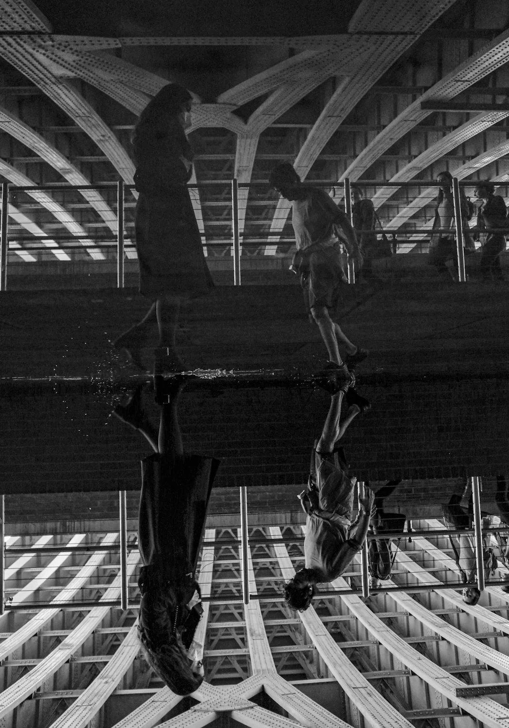 People standing on a wet surface with an architectural ceiling, reflected in a puddle on the ground, in black and white.