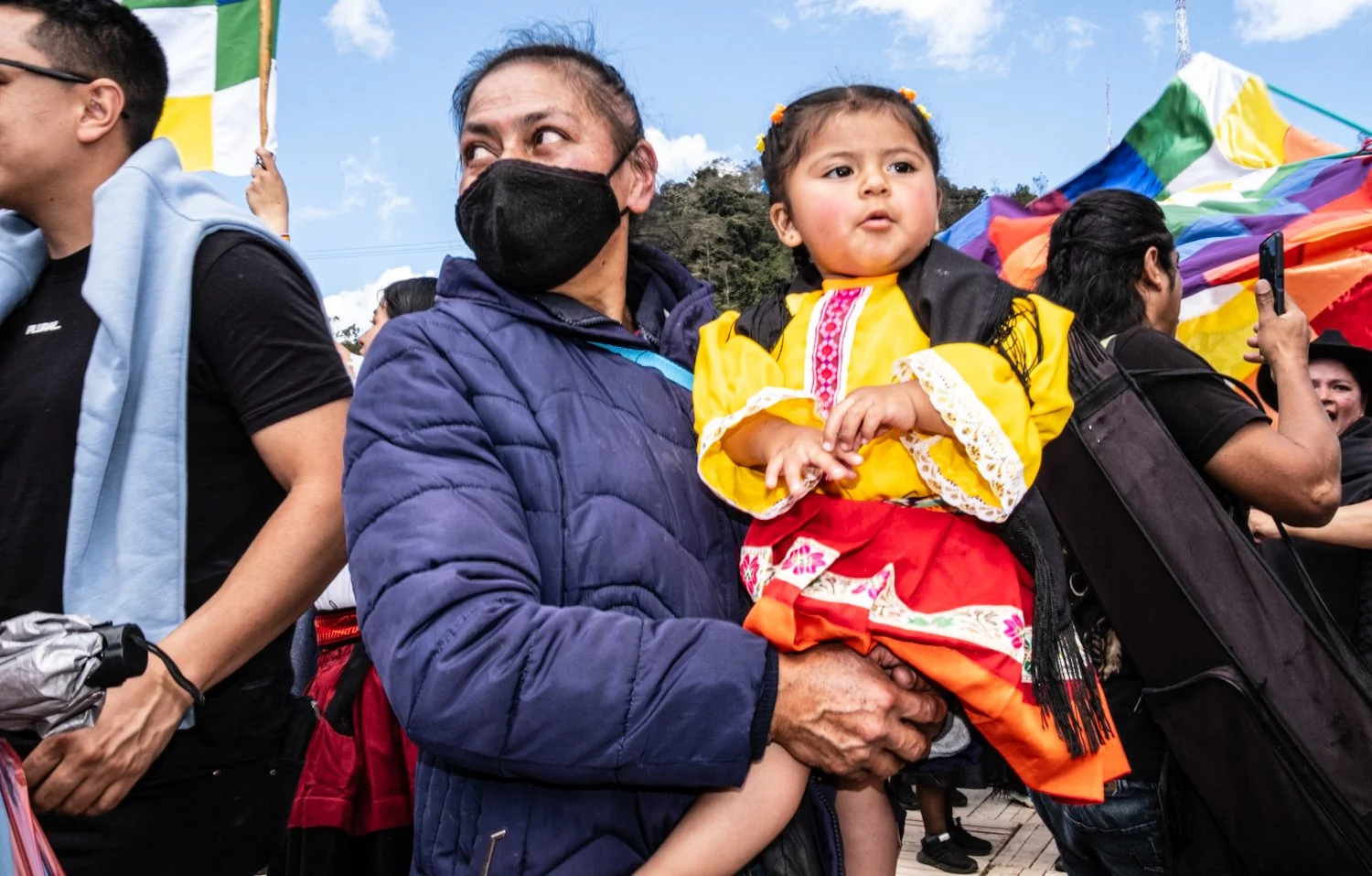 A man wearing a black face mask and a blue jacket holding a young girl in traditional Mexican clothing during an outdoor event with a crowd and rainbow flags.