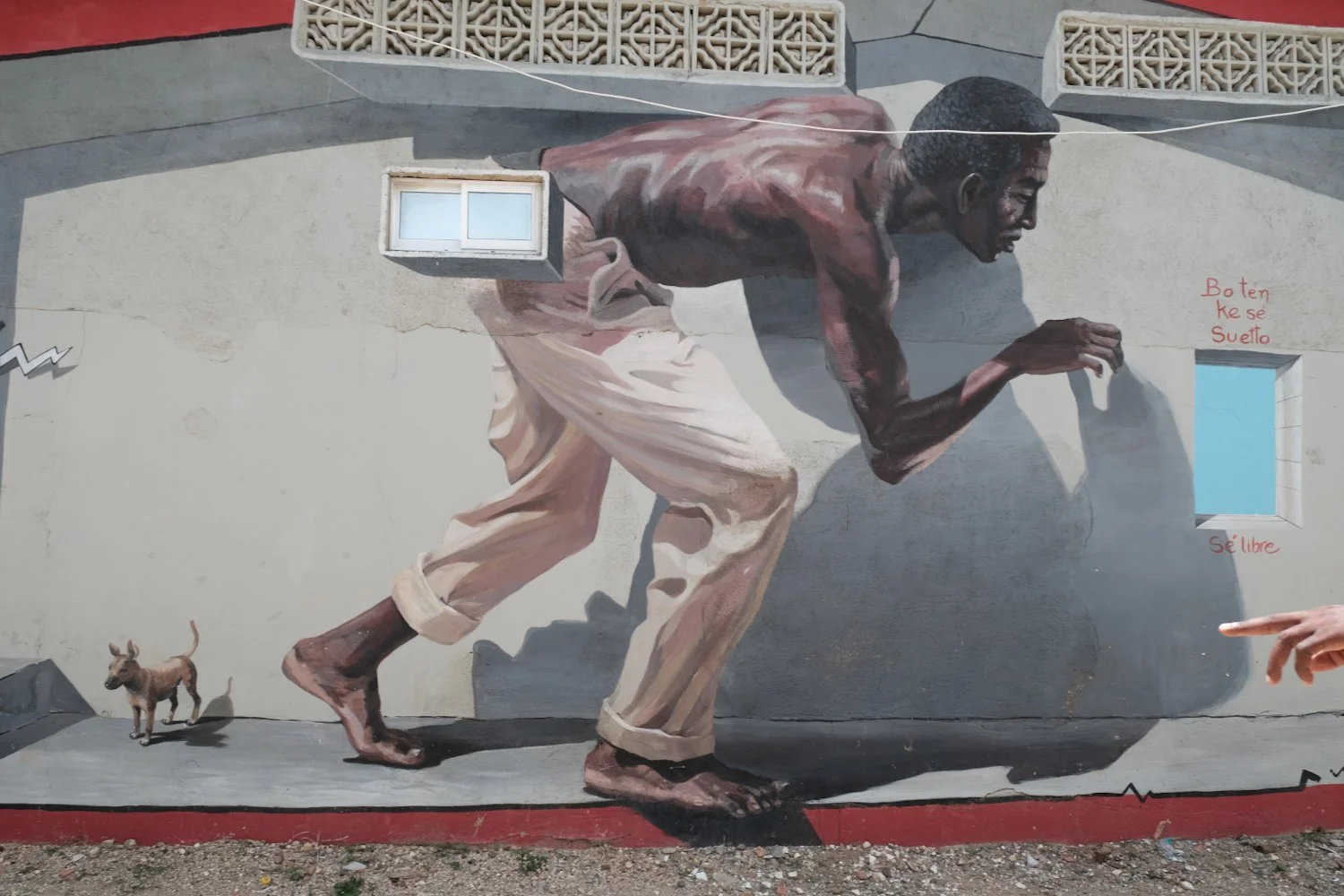 Street mural of a man with dark skin, short hair, and a muscular build, crawling forward on hands and feet, wearing loose beige pants. A small brown dog stands nearby, casting a shadow. Two small windows and a decorative concrete block wall are in th