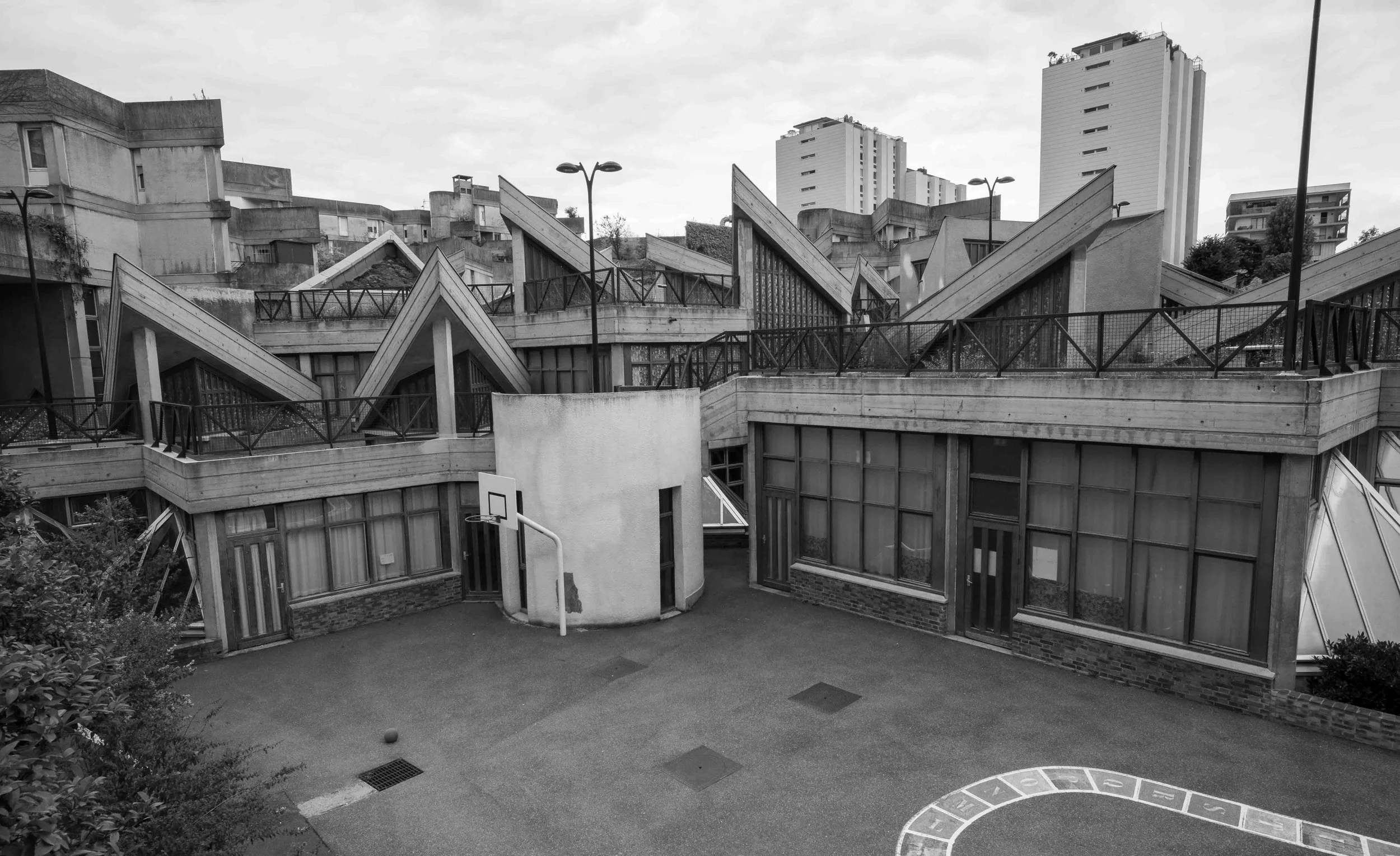Black and white photo of a modern concrete building with angular rooftops, surrounded by a small playground with a basketball hoop and hopscotch court, with high-rise buildings in the background.