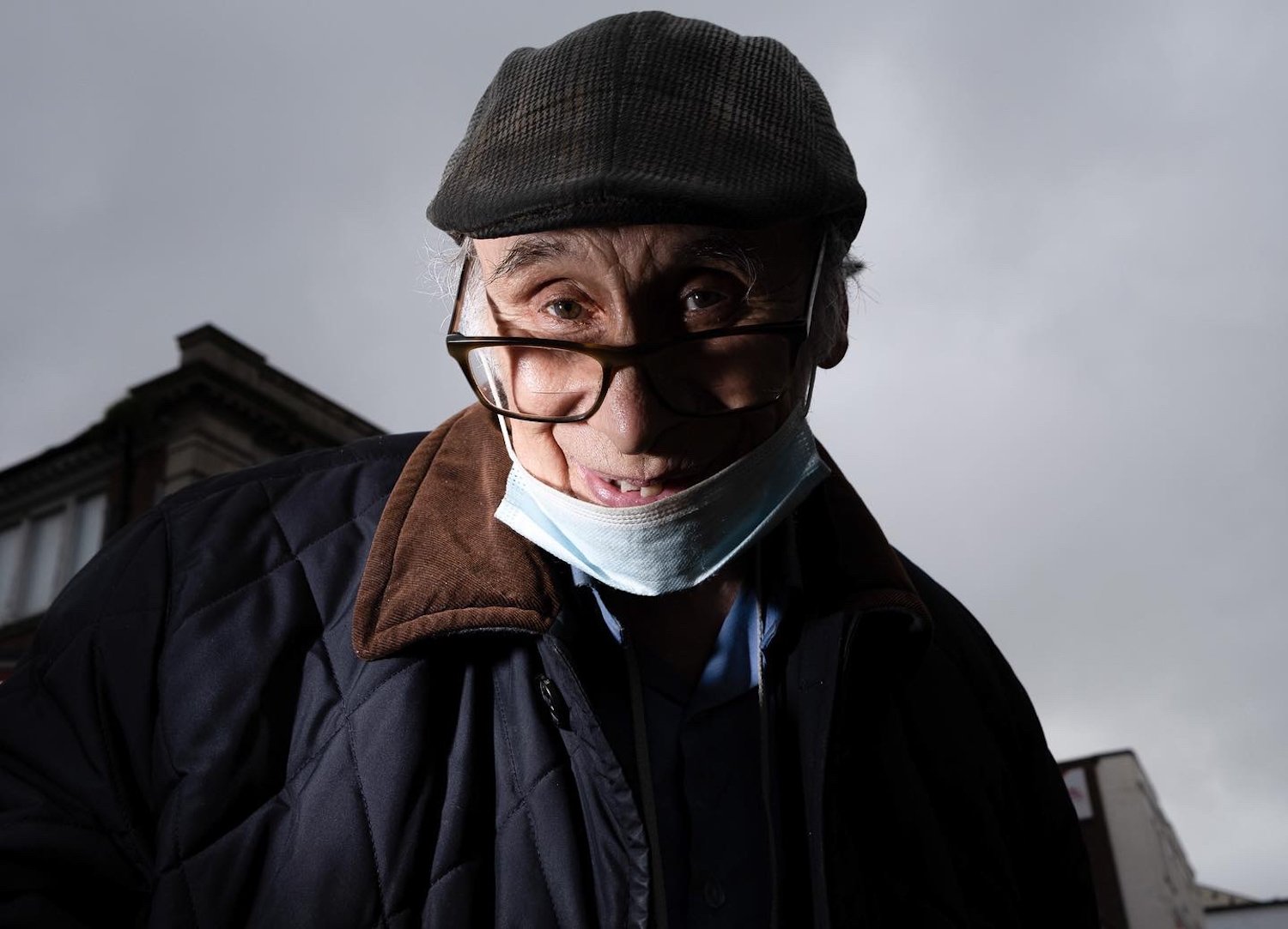 Close-up of an elderly man with glasses and a face mask lowered below his mouth, wearing a cap and coat, looking towards the camera with an expressive face, in an outdoor urban setting.