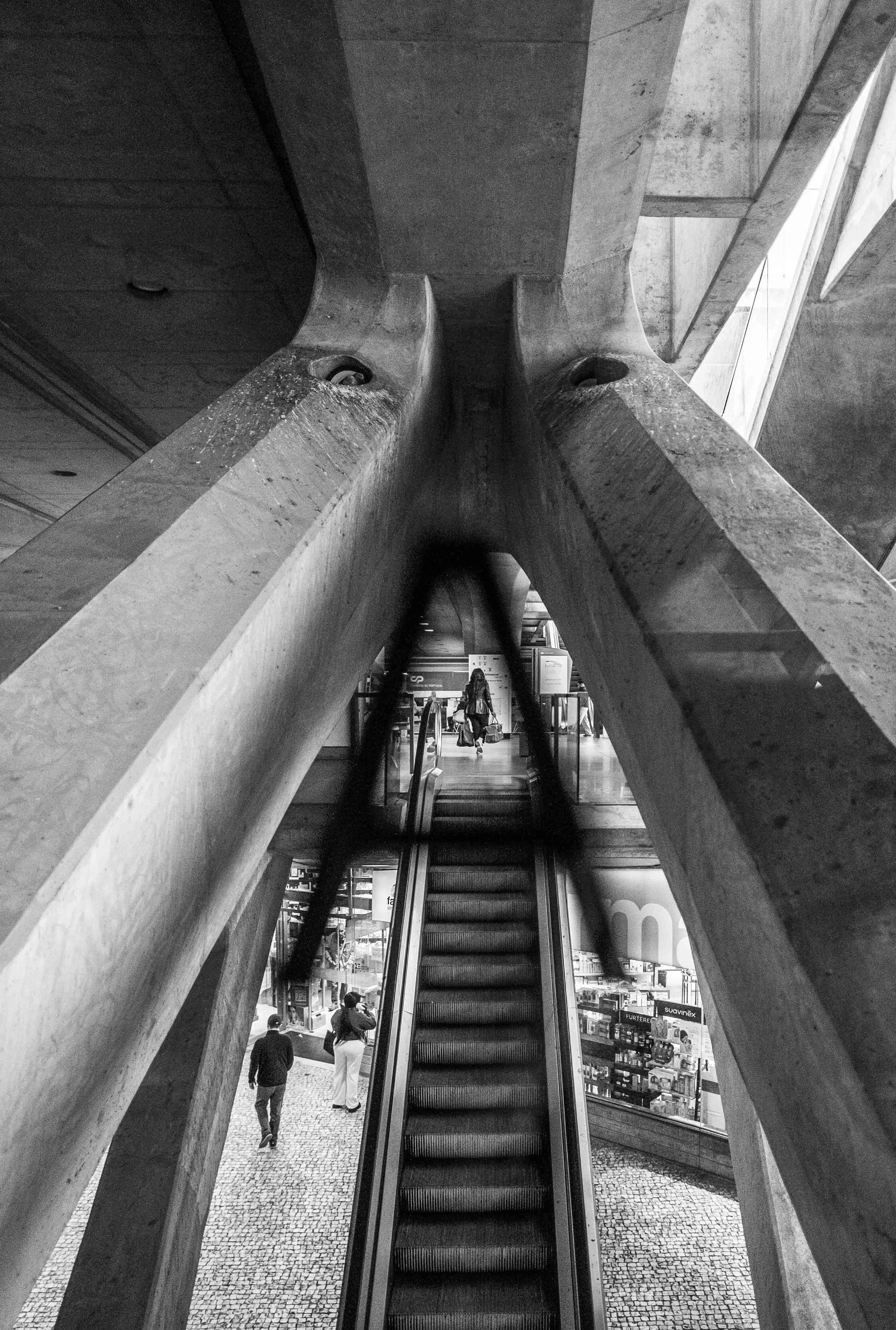 Black and white photo taken from below of a large concrete structure with a wide, straight escalator passing through it. People are seen walking and standing around in the background, which appears to be a shopping mall or similar commercial area.