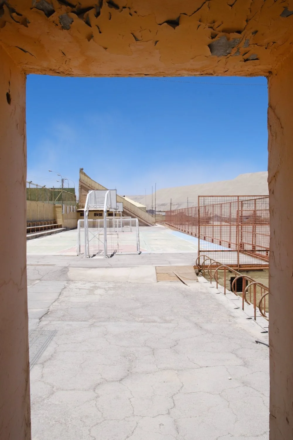 An outdoor basketball court with a hoop, a soccer goal, and a sandy desert in the background, viewed through a stone archway.