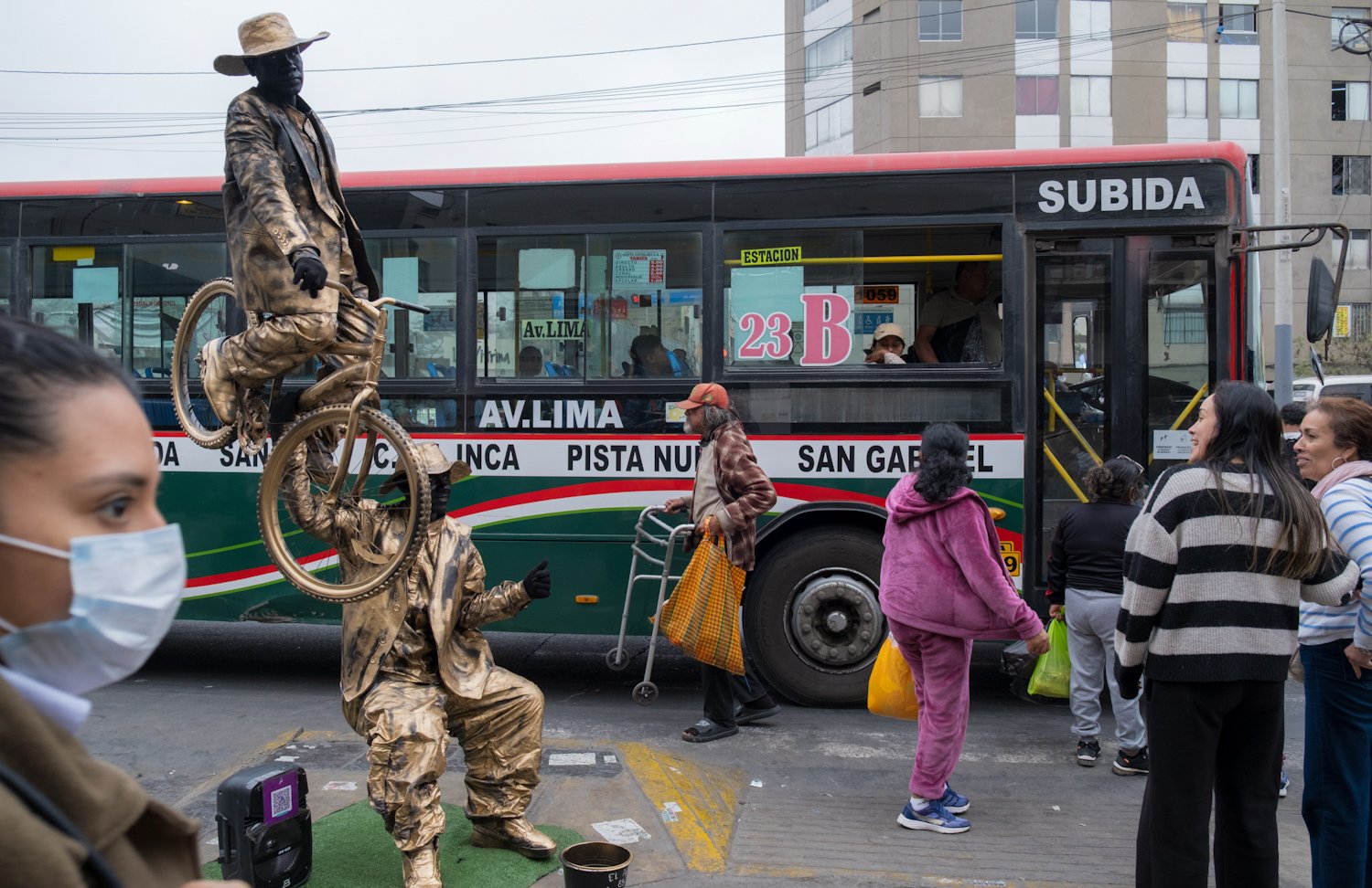 Street scene with a bus in the background and a street performer dressed as a golden statue in the foreground. The performer is standing on a platform, while another performer in a similar costume is crouching. Several pedestrians walk past, some wea