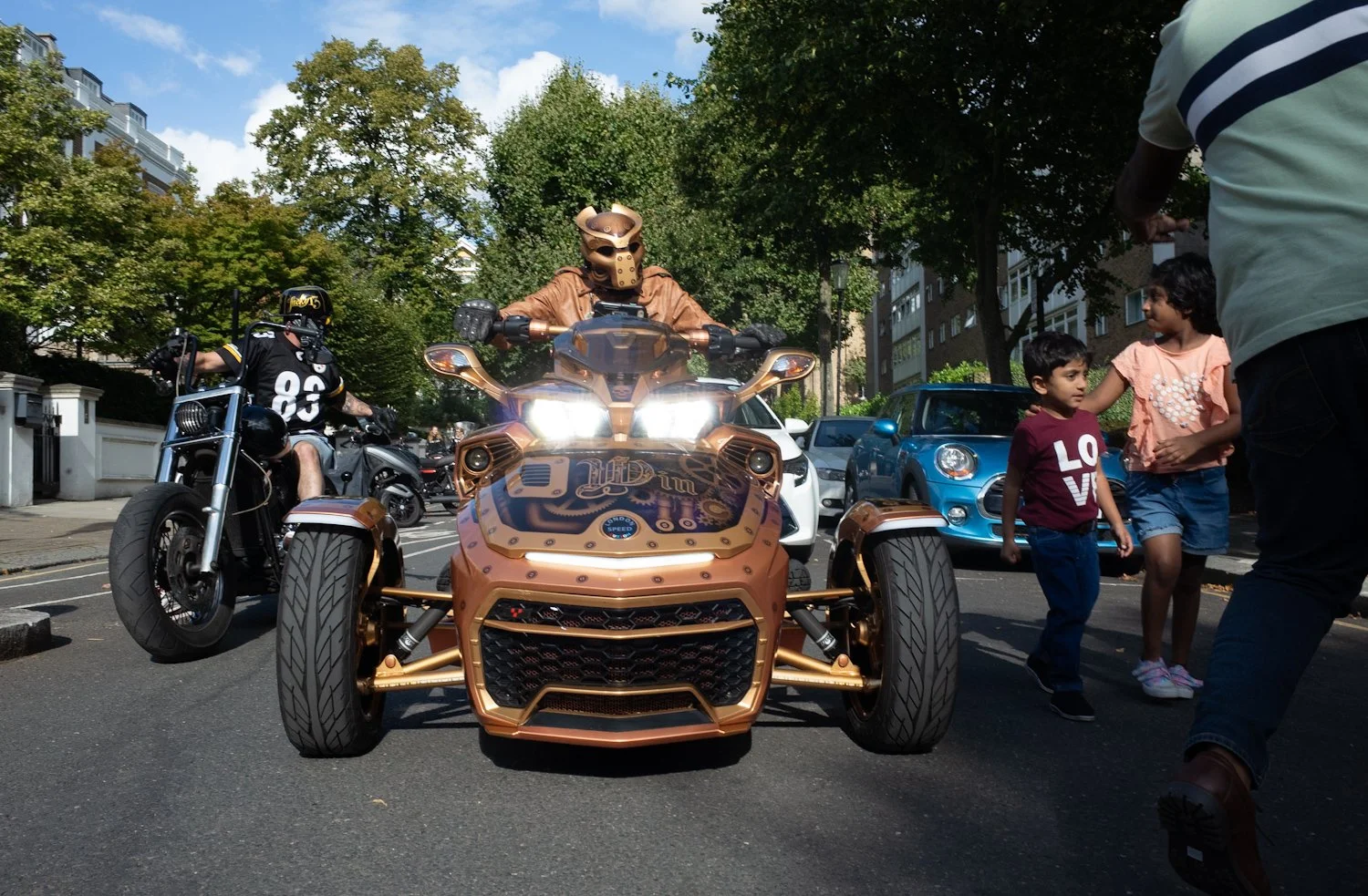 Man in bronze helmet riding a three-wheeled motorcycle, with a police officer on a black motorcycle beside him, children on the sidewalk, and trees and buildings in the background.