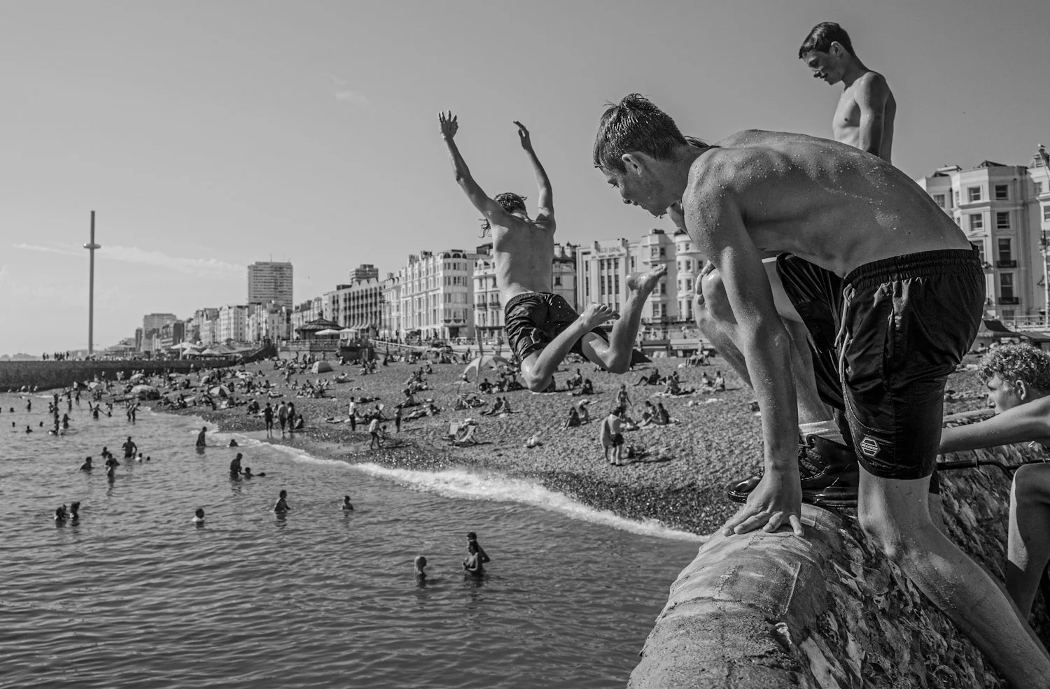 Children jumping into the water from a rock wall at the beach, with many people swimming and relaxing on the shore, and residential buildings along the coastline in the background.